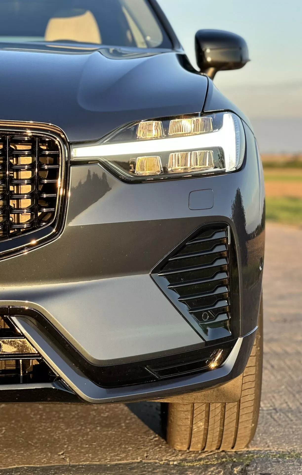 Pogled od blizu na sprednji levi del Volvo XC60 Recharge Plug-In Hybrid 2021, ki poudarja njegov navpični žaromet, LED žaromete z dnevnimi lučmi, stilske zračnike in meglenke, športen pokrov motorja in blatnike, kovinsko barvo s kontrastnimi obrobami in delček njegovega robustnega kolesa in pnevmatike, poudarjen z visokim sijajem vozila.