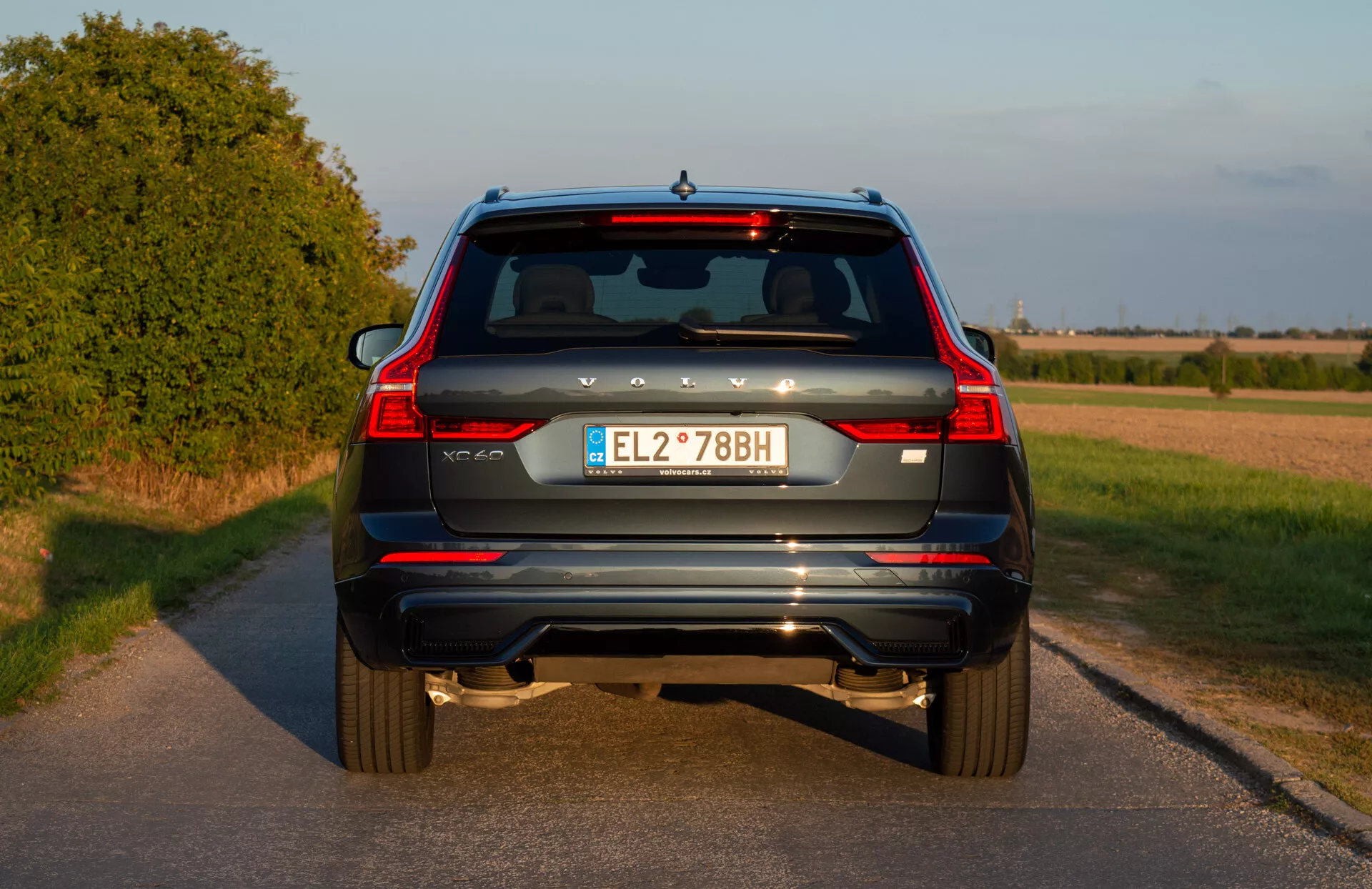 Zadnji pogled na modri SUV Volvo XC60 Recharge Plug-In Hybrid letnik 2021 z vertikalnimi zadnjimi lučmi, dvojnimi nasveti izpuha, zadnjimi reflektorji, brisalcem zadnjega okna, tretjo zavorno lučjo, evropsko registrsko tablico in zadnjo kamero na podeželski cesti pred sončnim zahodom