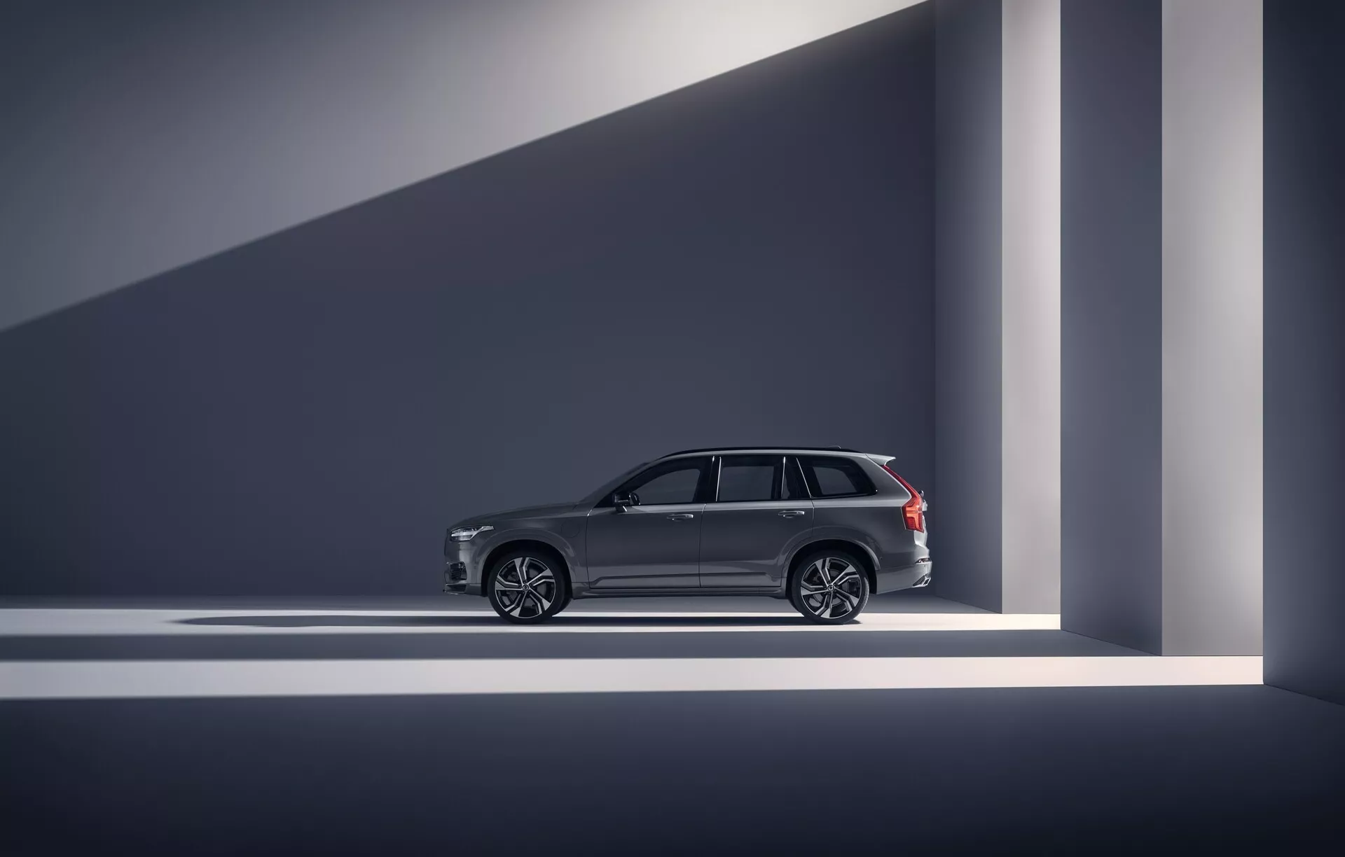 Stranski profil Volvo XC90 2015 (2. generacija) v kategoriji luksuznih SUV-jev. Fotografija je posneta v notranjem studiu ali razstavnem prostoru, prikazuje oblikovalske linije vozila, elegantne ročaje vrat, velika večkraka aluminijasta platišča in kromirane obrobe oken. Strešni nosilci nakazujejo uporabnost poleg luksuza. Vozilo je dobro osvetljeno z dramatična osvetlitvijo, ki ustvarja geometrične sence in poudarja njegov sodoben, visoko kakovosten videz. Napredne funkcije, kot je brezključni vstop, so nakazane s oblikovalskimi elementi.