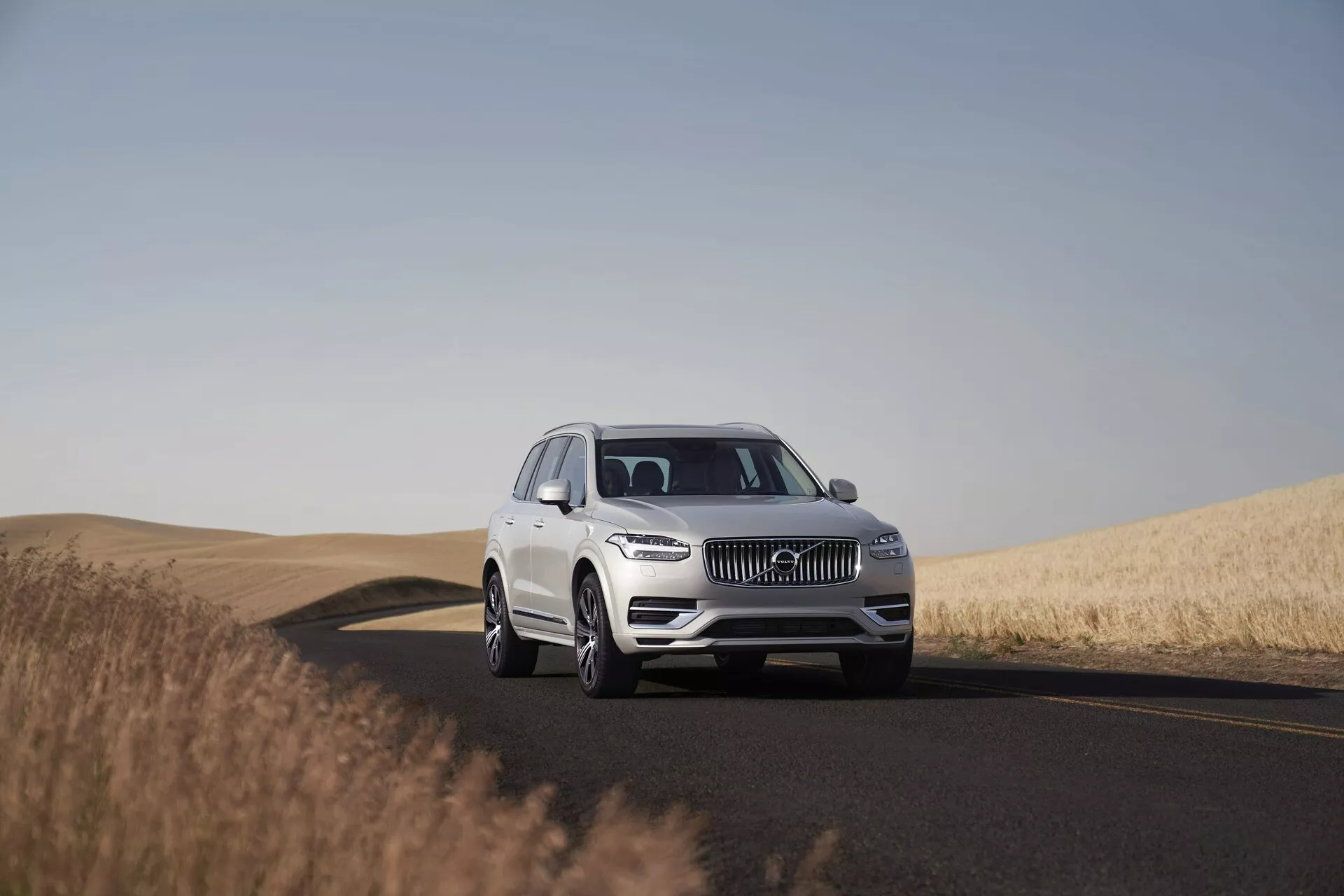 Beli SUV Volvo XC90 iz leta 2015 na dvopasovni cesti, posnet čez dan z jasnim nebom. Slika prikazuje vozilo iz nizkega do srednjega kota, kar poudarja njegovo mogočno prisotnost in dimenzije. Postavljen v središču okvirja s tretjinskim pogledom, fotografija zajame oblikovalske podrobnosti, kot so rešetka, LED žarometi, emblem in sprednji odbijač, kar kaže na napredne sisteme za pomoč vozniku. Kolesa, zlitine in morda velikost pnevmatik so jasno vidni. Mirno okolje predstavlja cesto, ki zavija v desno, in zlate pšenične poljance, kar poudarja primernost vozila za dolga potovanja v različnih pokrajinah.