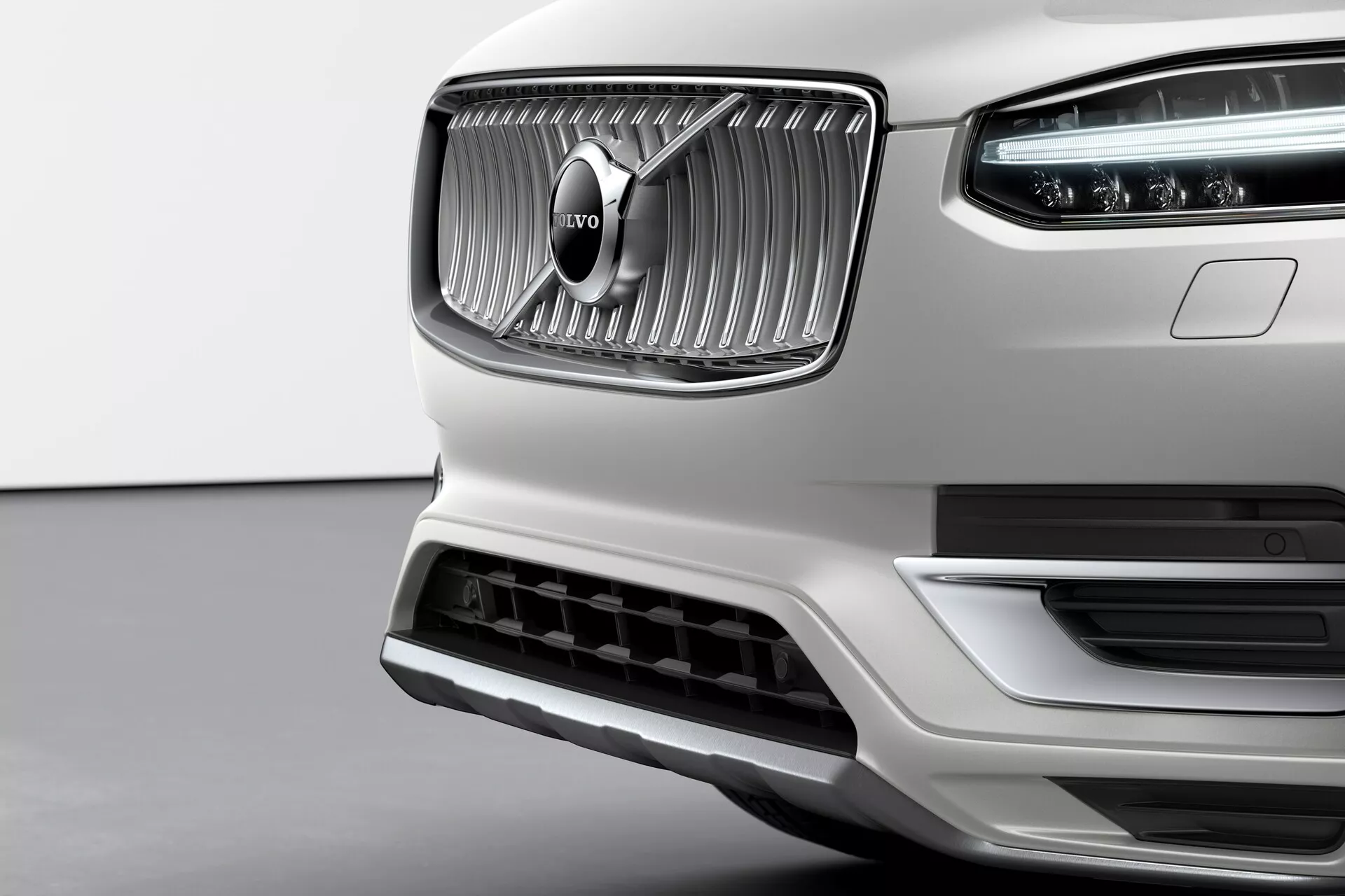 Blizu pogled prednjega dela belega Volvo XC90 druge generacije 2015, ki poudarja njegovo izrazito mrežo z navpičnimi letvami in značilnim Volvo emblemom v sredini. Kromirana površina mreže kontrastira z mat teksturo okoli nje. Vidne so LED dnevne luči Thorovo kladivo v glavnem žarometu. Spodnji odbijač ima velik vpis zraka s srebrnim robom, kar nakazuje na aerodinamičnost in hlajenje motorja. Posnetek iz rahlo nižjega kota poudarja eleganco, kakovost izdelave in premijski sprednji del dizajna Volvo.