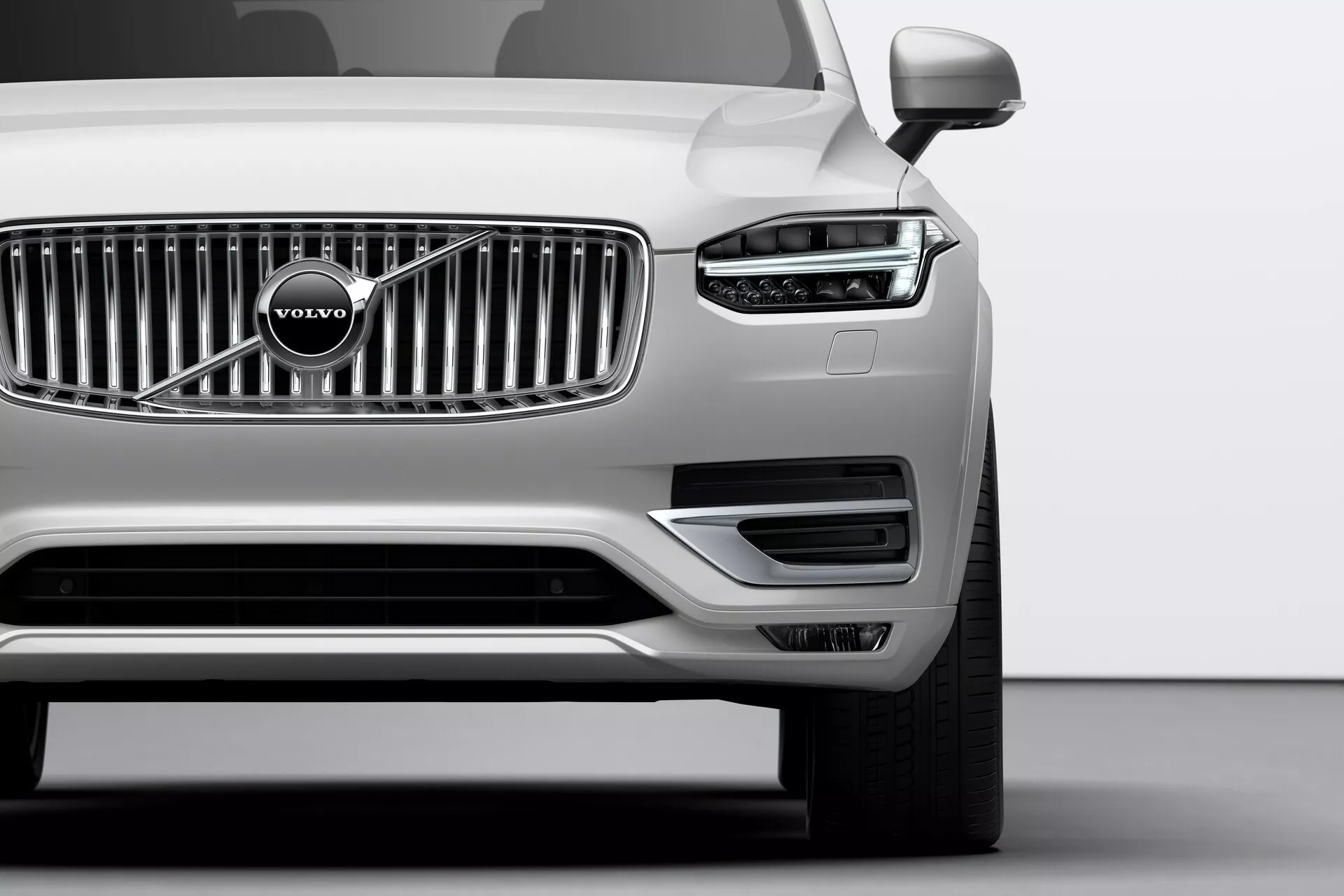 Posnetek od blizu prikazuje prednji del srebrnega Volvo XC90 iz leta 2015 (2. generacija) iz nizkega kota. Izpostavljena je izrazita kromirana mreža z značilnim emblemom Volvo, elegantni žarometi z LED dnevnimi lučmi v obliki črke T, imenovani 'Thorovo kladivo', ter dobro integrirani parkirni senzorji na sprednjem odbijaču. Spodnji del odbijača vsebuje zračnike in ohišja meglenk, obrobljena s srebrnim robom. Vidna vzvratna ogledala nakazujejo na napredne tehnološke funkcionalnosti, kot so indikatorji smeri ali sistemi za opozarjanje na mrtvi kot.