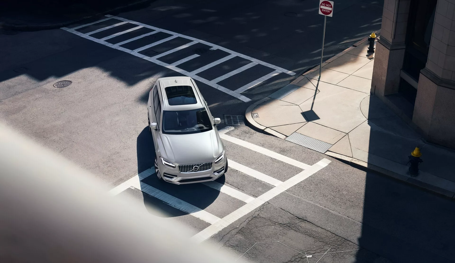 Posnetek s pticje perspektive Volvo XC90 iz leta 2015 (2. generacija) iz visokega kota, diagonalen pogled od sprednjega levega vogala; prikazuje svetel pokrov motorja, streho in del voznikove strani avtomobila. Vozilo je pravilno parkirano na označenem parkiranem mestu blizu prehoda za pešce in vsebuje oblikovne konture, ki nakazujejo aerodinamičnost, jasen vetrobran, ki razkriva delno viden notranji prostor, opazen strešni okno in ostre bočne linije. Mestno okolje poudarjajo bližnje oznake za parkiranje in prometne znake, z ostrimi sencami, ki nakazujejo poldnevno osvetlitev. Vozilo tudi nakazuje sodobne tehnološke funkcije brez vidnih senzorjev in kamer.