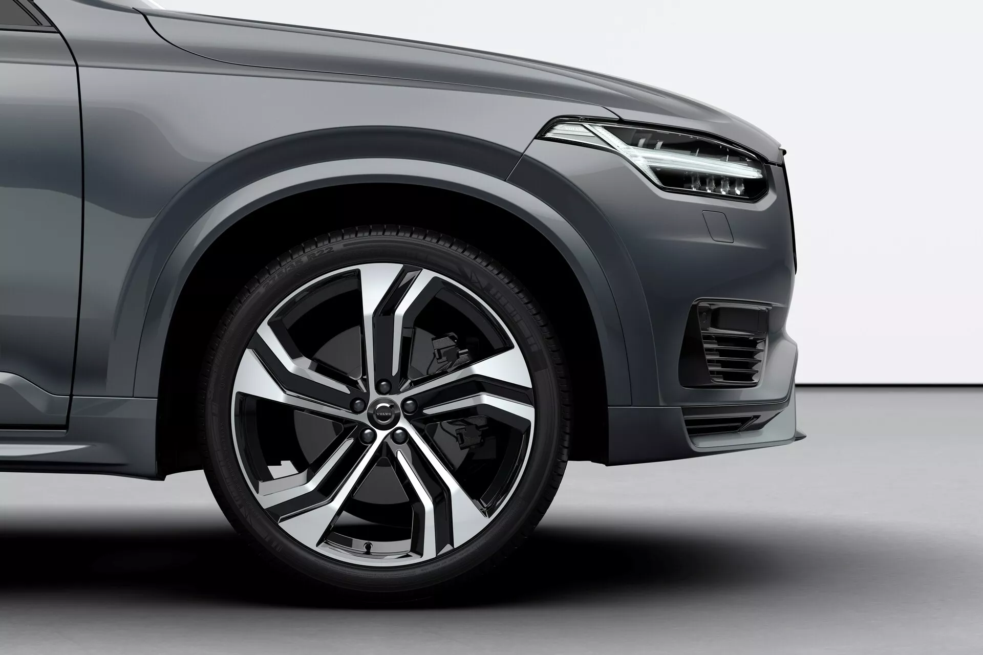 Nizkoten snemalni kot sprednjega kolesa na strani voznika in sosednjih delov vozila Volvo XC90 iz leta 2015, ki prikazuje sodoben dizajn aluminijskega platišča z več kraki, športno pnevmatiko z nizkim profilom, del prednjega blatnika z aerodinamičnimi linijami, eleganten dizajn žarometa, sprednji odbijač z vodoravnim vzorcem rešetke, možno meglenko ali zračnik pod žarometom in kovinski zaključek barve, kar vse skupaj nakazuje na luksuzno ali višji nivo opremljenosti vozila.