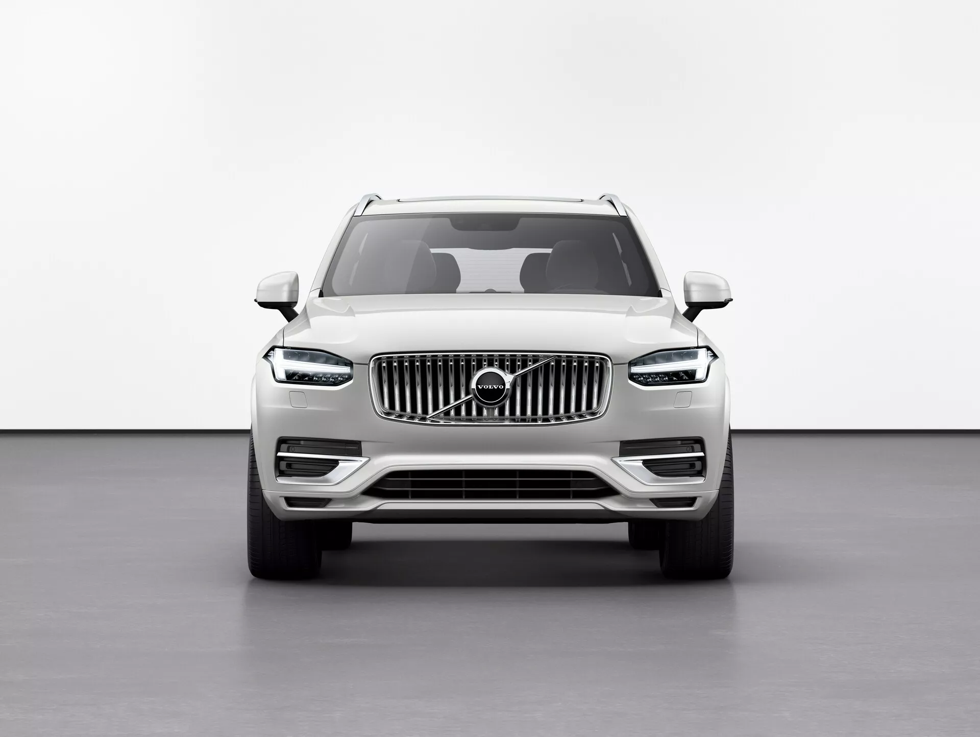Sprednji pogled na SUV Volvo XC90 2015 (2. generacija) pred belim ozadjem v studijskem okolju, ki poudarja povečano višino vožnje, veliko mrežo z navpičnimi letvami z emblemom Volvo, elegantne LED žaromete 'Thorovo kladivo', definirane linije pokrova motorja, meglenke na spodnjem odbijaču in stranska ogledala z vgrajenimi smerniki.