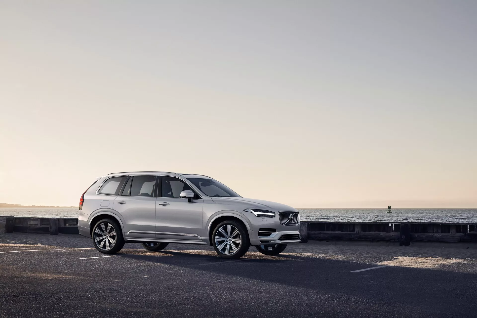 Stranski profil belega SUV Volvo XC90 2015 na obalni cesti, z vidnimi oblikovalskimi značilnostmi, vključno z obliko koles, vratnimi ploščami, stranskimi ogledali, strešnimi nosilci in konfiguracijo oken, poudarjenimi z mehko svetlobo ob zori ali mraku.