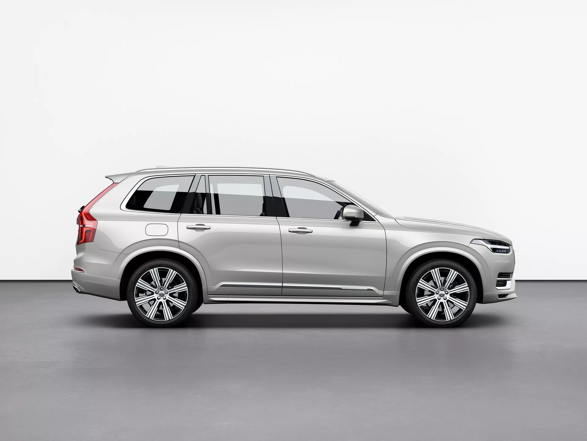Stranski profil srebrnega SUV Volvo XC90 2015 (2. generacija), ki prikazuje njegova lita platišča, strešne nosilce, stransko ogledalo z vgrajenim smernikom in podrobne karoserijske linije, umeščen centralno proti preprostemu ozadju.