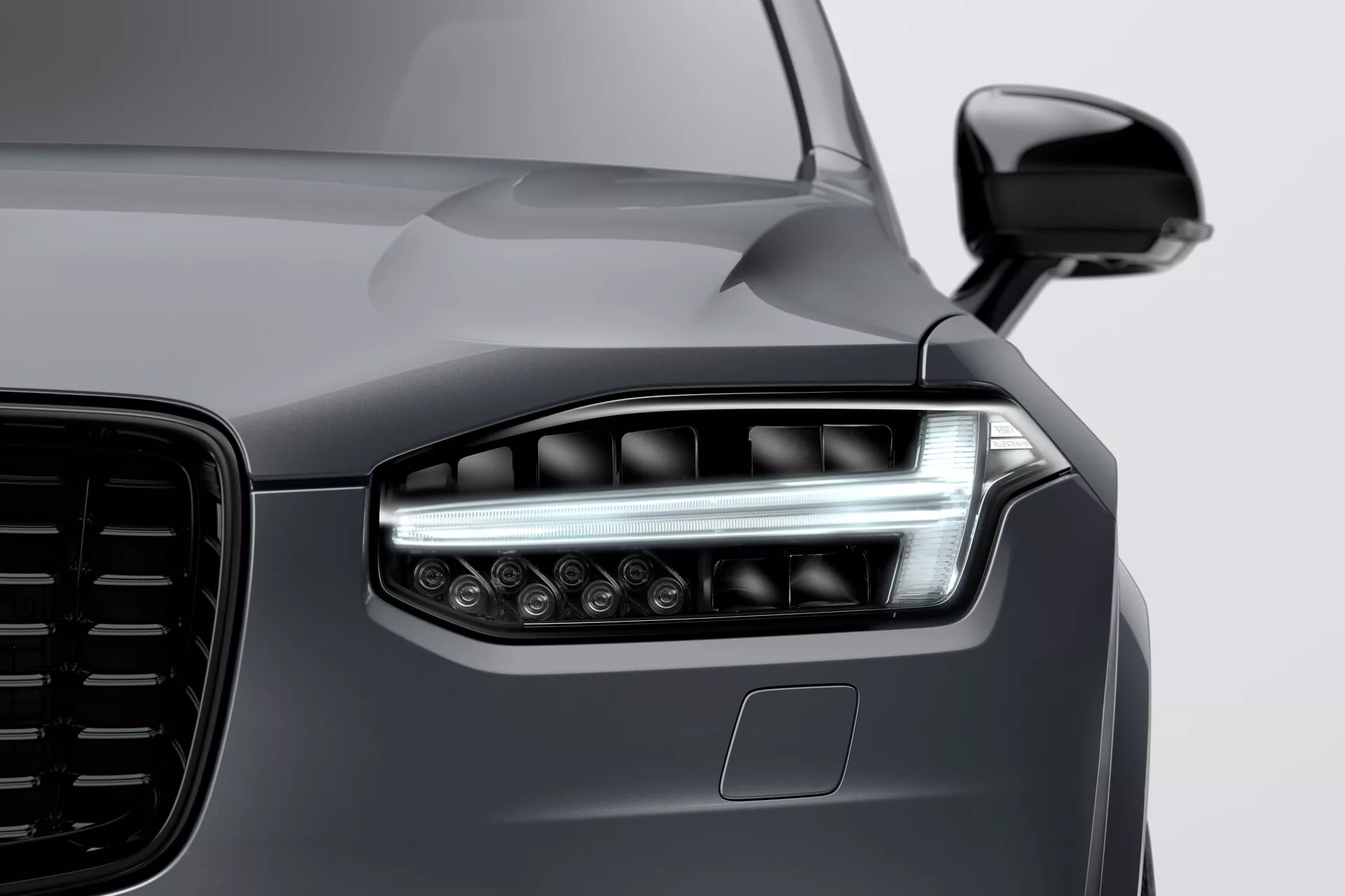 Povečan posnetek sprednjega konca Volvo XC90 2015 (2. generacija), ki poudarja levi žaromet z moderno LED ali lasersko tehnologijo, dvojnimi svetlobnimi elementi, elegantnimi dnevnimi lučmi (DRL) in več projekcijskimi lečami za boljšo vidljivost. Sliko poudarja velika estetsko oblikovana mreža, aerodinamično stransko ogledalo v barvi karoserije in morebitni ADAS senzor pod žarometom. Gladek, odsevujoč globok kovinsko siv zaključek poudarja elegantne, a mišičaste linije pokrova motorja in blata, kar kaže na visoko kakovostno barvanje in vrhunsko obliko vozila.