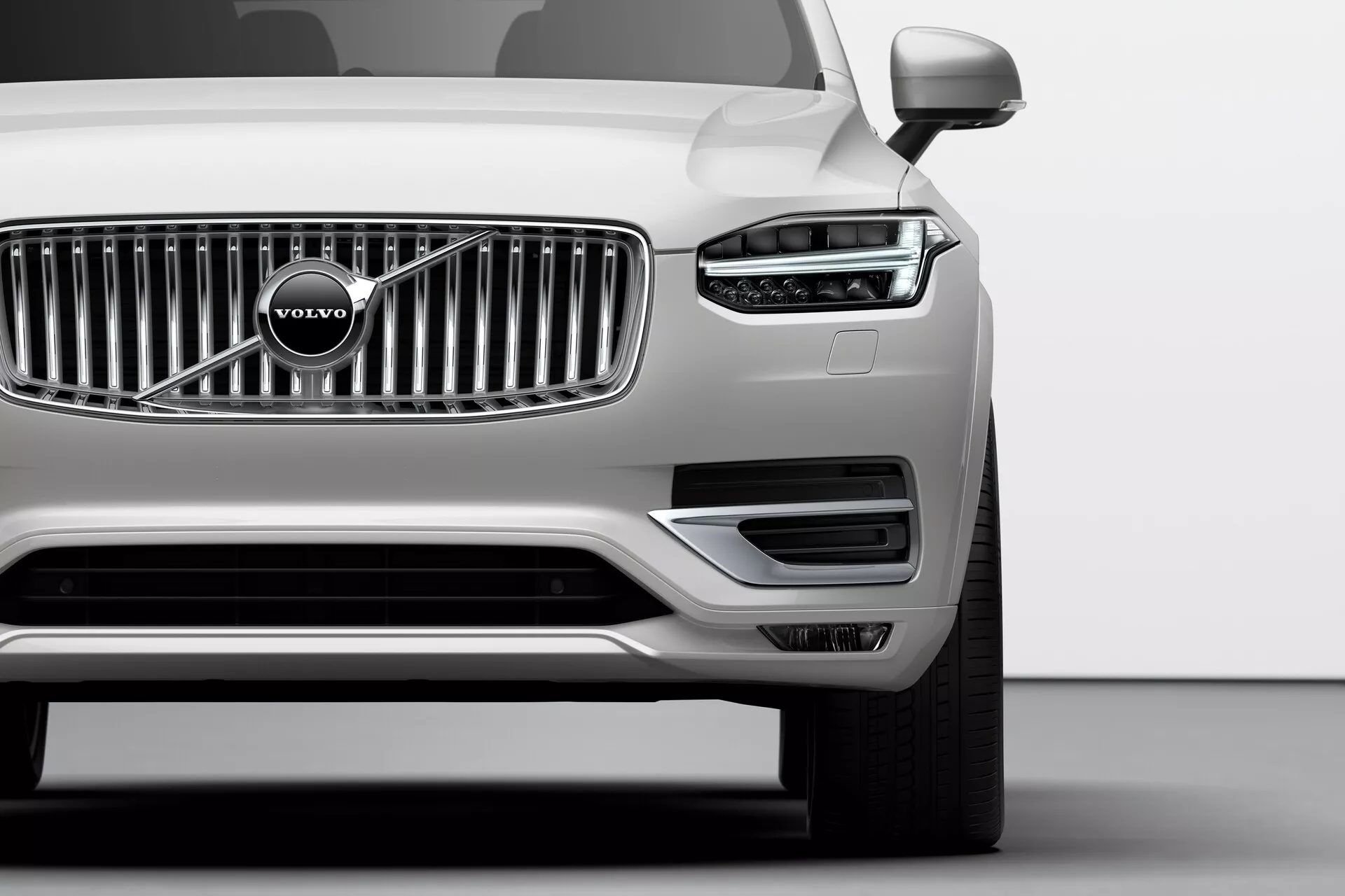 Fotografija srebrnega Volvo XC90 iz leta 2015 (2. generacija) iz nizkega kota v studiu, prikaz prednjega levega kota vozila, ki poudarja značilen LED vzorec dnevne luči v obliki Thorovega kladiva, vertikalno kromirano rešetko z Volvovim logom, minimalističen prednji odbijač z zračnimi režami, levo stransko ogledalo, večkrako platišče iz zlitin in brezhiben kovinski premaz, ponazarjajoč napredno svetlobno tehnologijo, oblikovanje blagovne znamke in aerodinamične lastnosti.