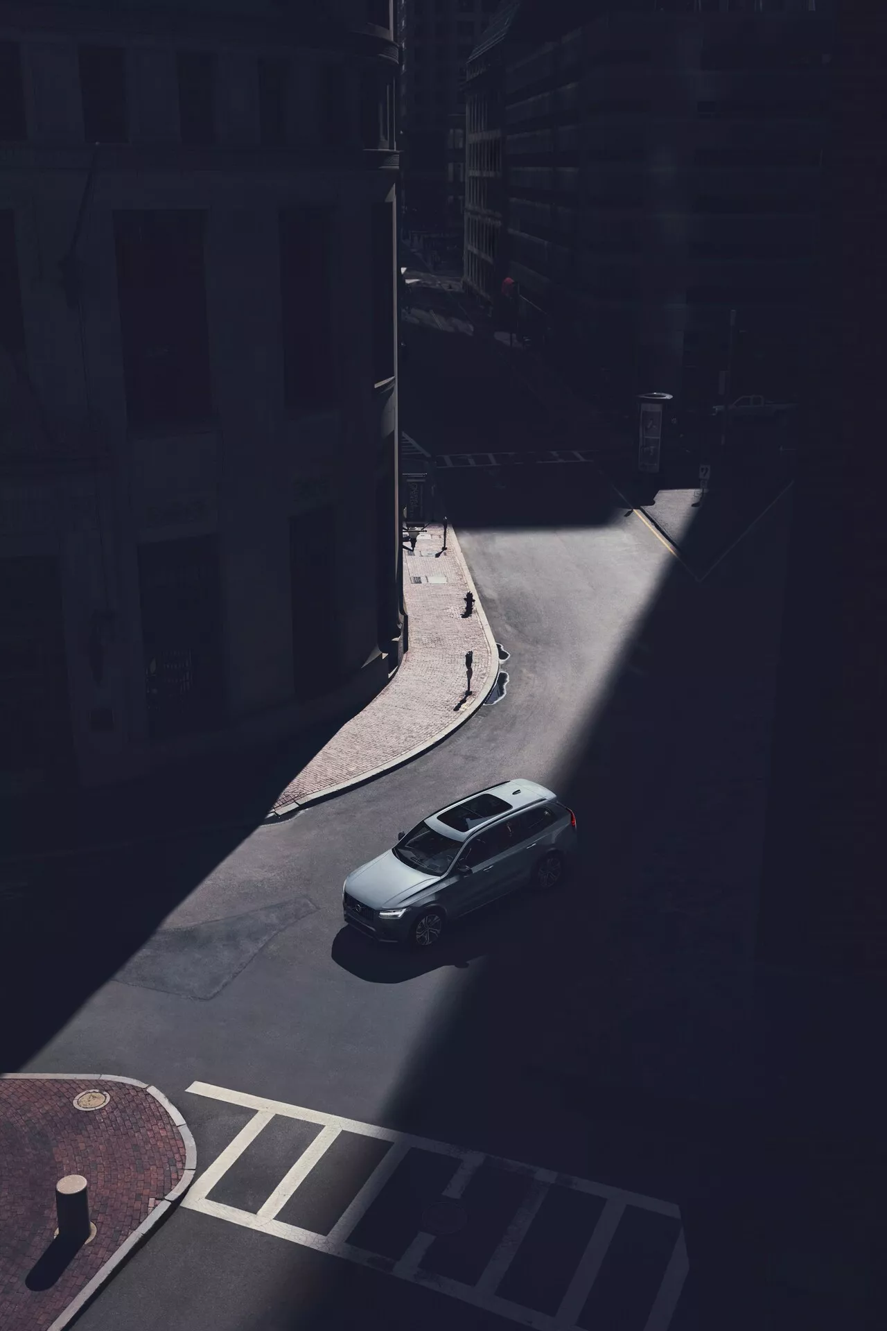 Zračni pogled na Volvo XC90 sedan iz leta 2015 (2. generacija), parkiran na senčnem mestnem cestišču, posnet iz visoke perspektive pod poševnim kotom. Jasno so vidni prtljažnik, streha in zadnje vetrobransko steklo avtomobila, pri čemer je zadnji del vozila osvetljen s sončno svetlobo. Slika prikazuje zadnji pokrov prtljažnika, obliko zadnjega okna in zgornji del prtljažnika, medtem ko sence zakrivajo specifične oblikovalske podrobnosti, kot so zadnje luči in odbijač. Gladka aerodinamična strešna linija je očitna, brez vidnih strešnih nosilcev ali strešnih oken. Avto je parkiran ob robniku blizu fasad stavb in prehoda za pešce, kar poudarja osamljenost vozila v senčnem mestnem okolju.