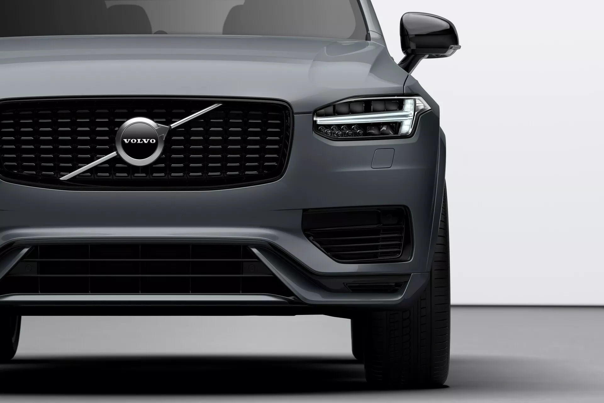 Bližnji pogled na sprednji kot Volvo XC90 2015 (druga generacija), osredotočen na levo stran, prikazuje značilno rešetko, velik emblem Volvo in zasnovo LED žarometov 'Thorovo kladivo'. Slika vključuje žaromet, sprednjo kolo, del voznikovega vrat, vidne senzorje na spodnjem odbijaču in stransko ogledalo z morebitno tehnologijo nadzora mrtvega kota.
