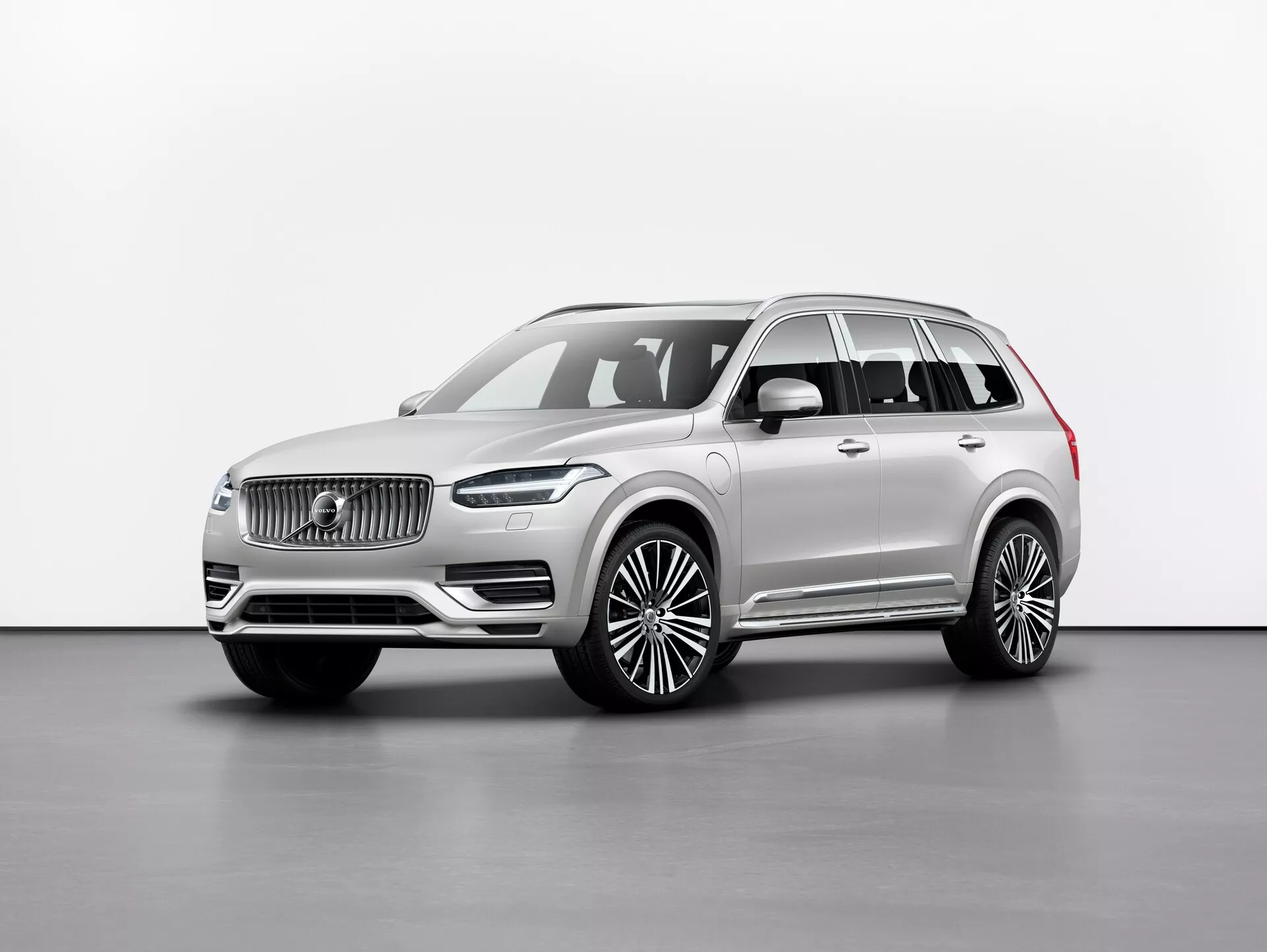 Studijska fotografija luksuznega SUV-ja Volvo XC90 2015 (2. generacija), prikazana iz tričetrtinskega sprednjega kota, prikazuje sprednjo masko z izrazitim logotipom, elegantne LED žaromete, velik sprednji odbijač in dolgo medosno razdaljo z velikimi litimi platišči. Bočna ogledala so nameščena pod stebričkom A s kromiranimi obrobami oken in strešnimi nosilci, ki prispevajo k občutku prestiža. Nevtralna barva laka izžareva prefinjenost, slika pa poudarja linije in konture avtomobila z enakomerno osvetlitvijo na preprostem ozadju. Verjetno vključuje tehnologije za asistenco vozniku in napredne informacijsko-zabavne sisteme.