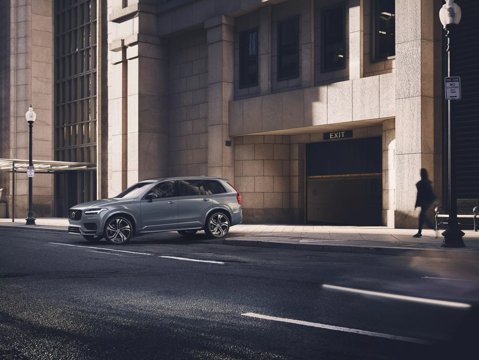 Moderni SUV Volvo XC90 2015 (2. generacija) parkirano na mestni ulici, posneto iz perspektivnega kota, ki prikazuje stranski profil in del sprednje maske. Lesketajoča kovinska barva vozila poudarja njegovo dobro vzdrževano stanje, večkraka aluminijasta platišča in zatemnjena okna. Slika je posneta z ravni oči povprečnega odraslega, v ospredju prikazuje avtomobil, ki ga obdajajo mestni elementi, vključno s fasado stavbe, silhueto pešca in ulično svetilko v zgodnjem jutranjem ali pozno popoldanskem soncu.
