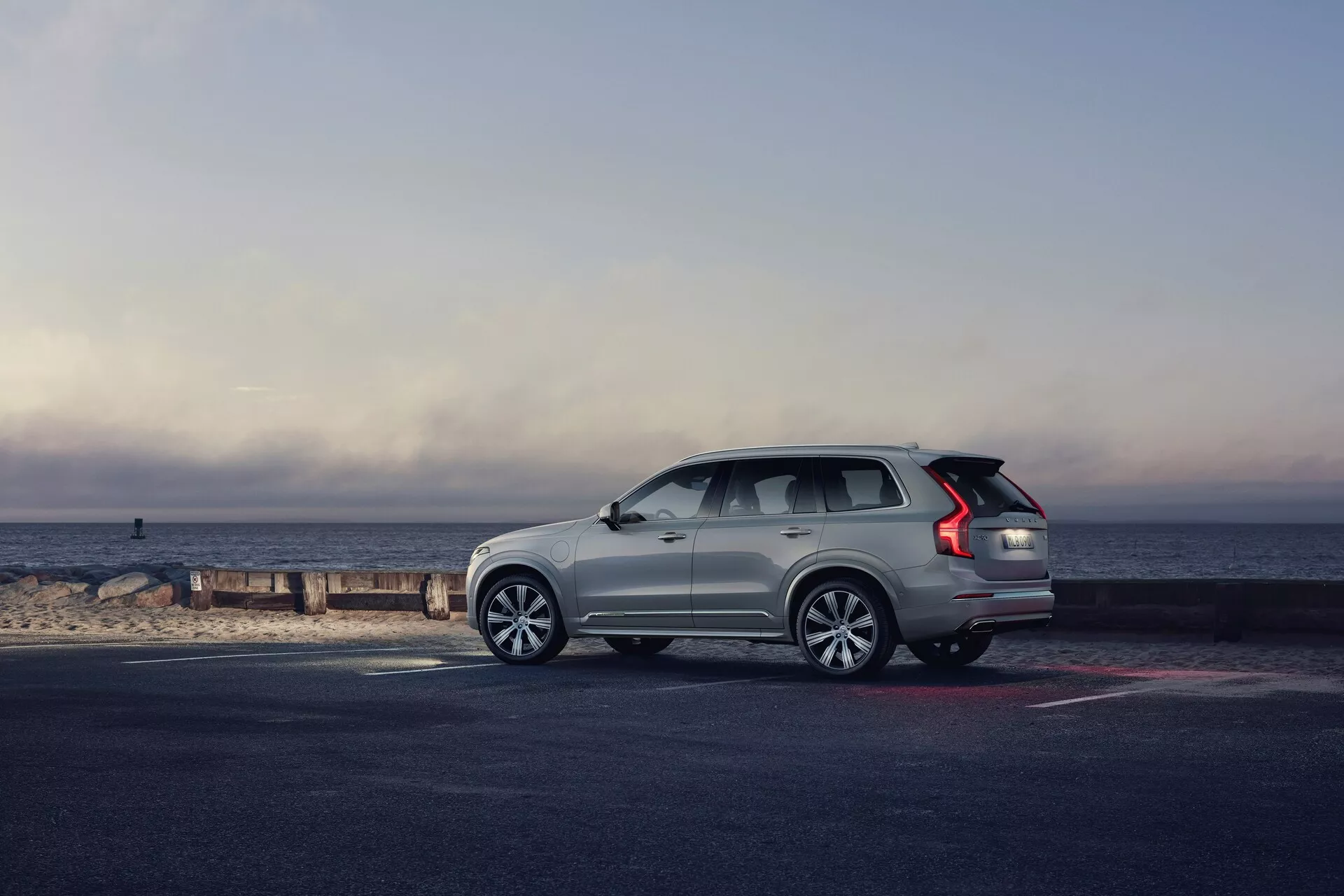 Profilni pogled na Volvo XC90 SUV iz leta 2015 (2. generacija), parkiran ob obali, s vozilom, obrnjenim v desno. Nagib poudarja stran in zadnji del, prikazuje elemente oblikovanja, kot so večkrake aluminijaste platišča, podaljšane LED zadnje luči, strešni nosilci, naraščajoča linija oken in ročaji vrat na ravni s karoserijo. Svetlo obarvan SUV izstopa na temnejšem pločniku in plaži, osvetljen z naravno jutranjo ali pozno popoldansko svetlobo, s slikovitim ozadjem obzorja, kjer se nebo sreča z morjem. Celotna kompozicija vzbuja občutek avanture, razkošja in svobode.