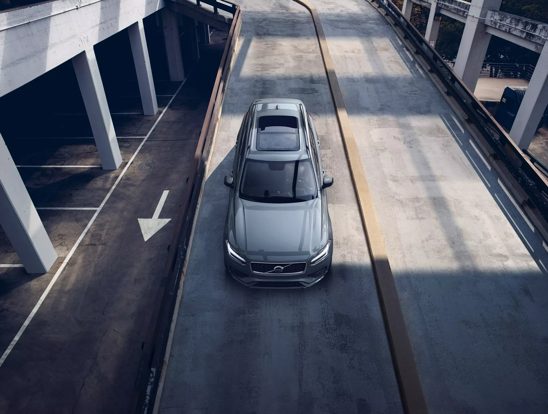 Pogled iz ptičje perspektive na siv ali srebrn Volvo XC90 sedan iz leta 2015, ki potuje po večnivojski mestni cesti, prikazuje panoramsko streho, aerodinamično zasnovo, elegantne LED žaromete in velika aluminijasta platišča, kar nakazuje sodobno luksuzno vozilo.