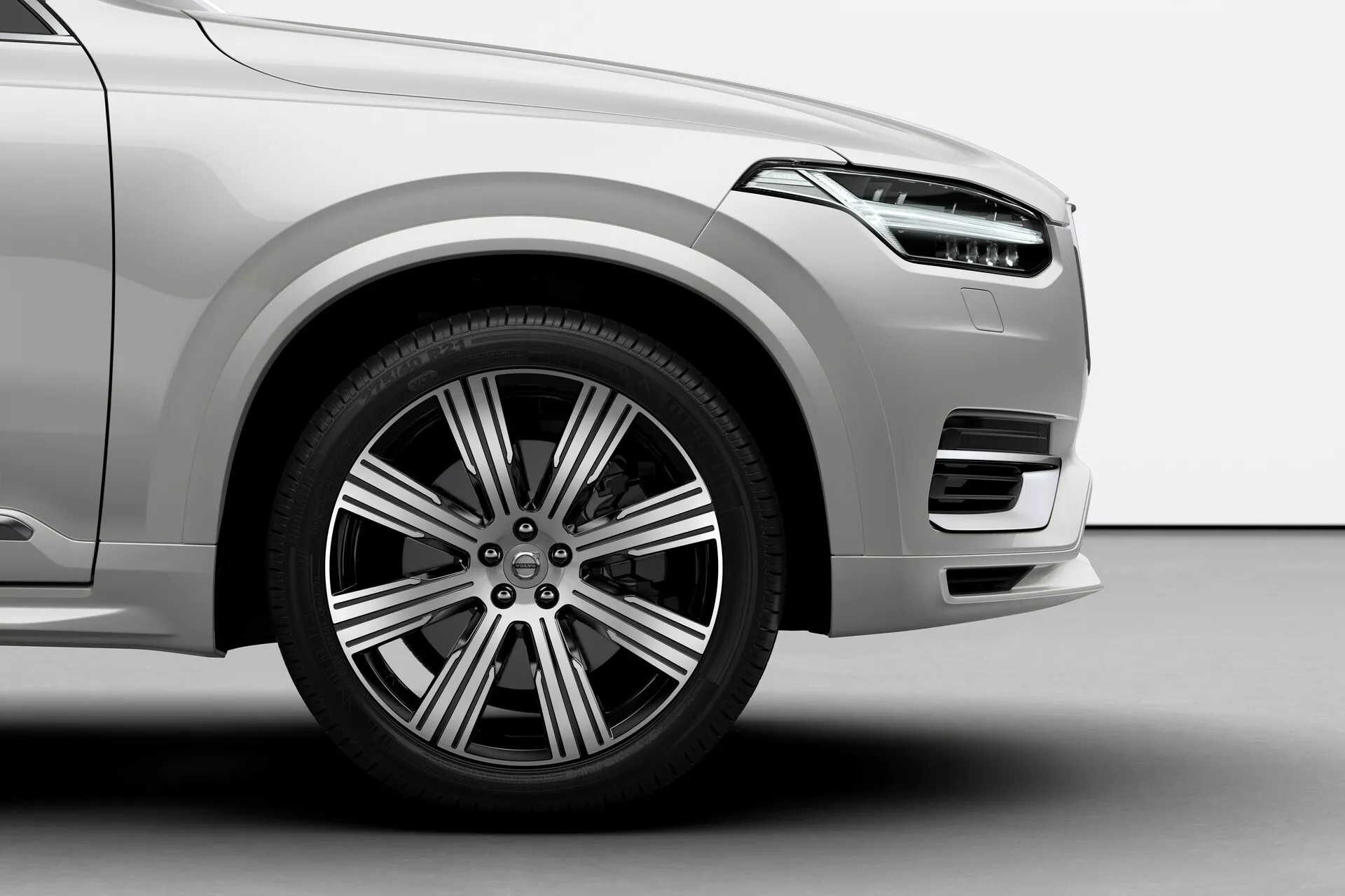 Od blizu, posnetek z nizkega kota prednje četrtpanelke vozila Volvo XC90 2015 2. generacije, ki prikazuje prednje kolo na strani voznika z večkrako zasnovo iz lahke zlitine, zavorni sistem z diski, viden zavorni cilinder in cestno pnevmatiko. Slika poudarja prednji odbijač z velikim vnosom zraka, svetlo kovinsko barvo ter gladke zaobljene aerodinamične linije.