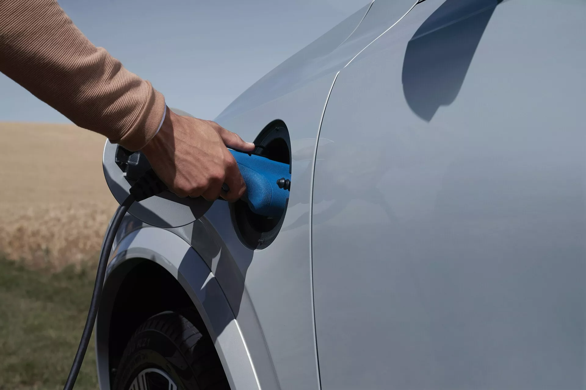 Stranski pogled na električno ali plug-in hibridno vozilo Volvo XC90 2015 druge generacije med polnjenjem. Posnetek zajema stran vozila s poudarkom na polnilni vtičnici in vtiču, delno prikazuje zadnje kolo in spodnji del avtomobila. Povezan modri polnilni kabel, ozadje odprtega naravnega območja, kar nakazuje podeželsko okolje.