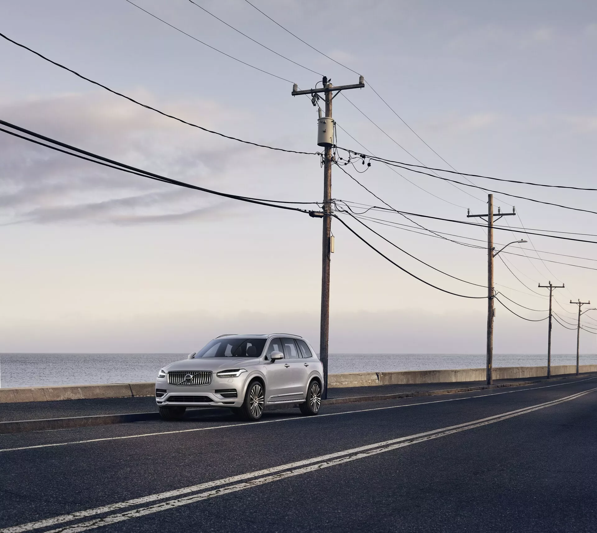 Volvo XC90 druge generacije iz leta 2015, luksuzni SUV parkiran na asfaltni cesti vzporedni z oceansko obalo, fotografirano pri dnevni svetlobi z mehko razpršeno svetlobo in dolgimi sencami. Pogled iz sprednjega kota poudarja elegantne oblikovne elemente, vključno s žarometi, mrežo in obrisi pokrova motorja, svetlo srebrna ali bledo šampanjska barva kontrastira s temno cesto in naravnim ozadjem.