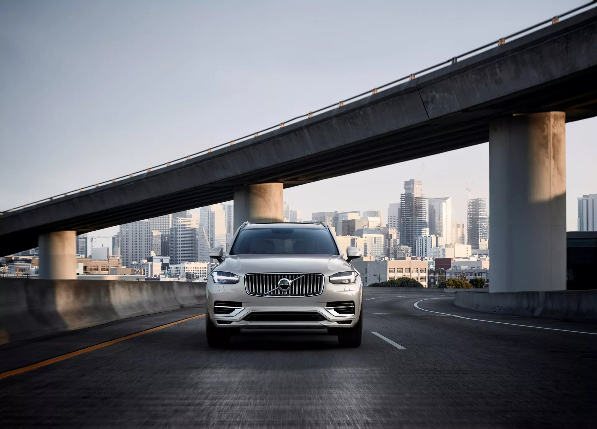 Frontalni posnetek SUV Volvo XC90 druge generacije iz leta 2015 na praznem nadvozu, s panoramo mesta v ozadju. Kot kamere je nizek, kar poudarja sprednjo masko, LED žaromete, kromirane poudarke, lita kolesa in oblikovan odbijač s senzorji, ki kažejo na pomoč pri parkiranju ali sistem za izogibanje trkom. Perla ali kovinska barva avtomobila se sveti v dnevni svetlobi, poudarjajoč njegov luksuz in sodoben dizajn v urbanem okolju.