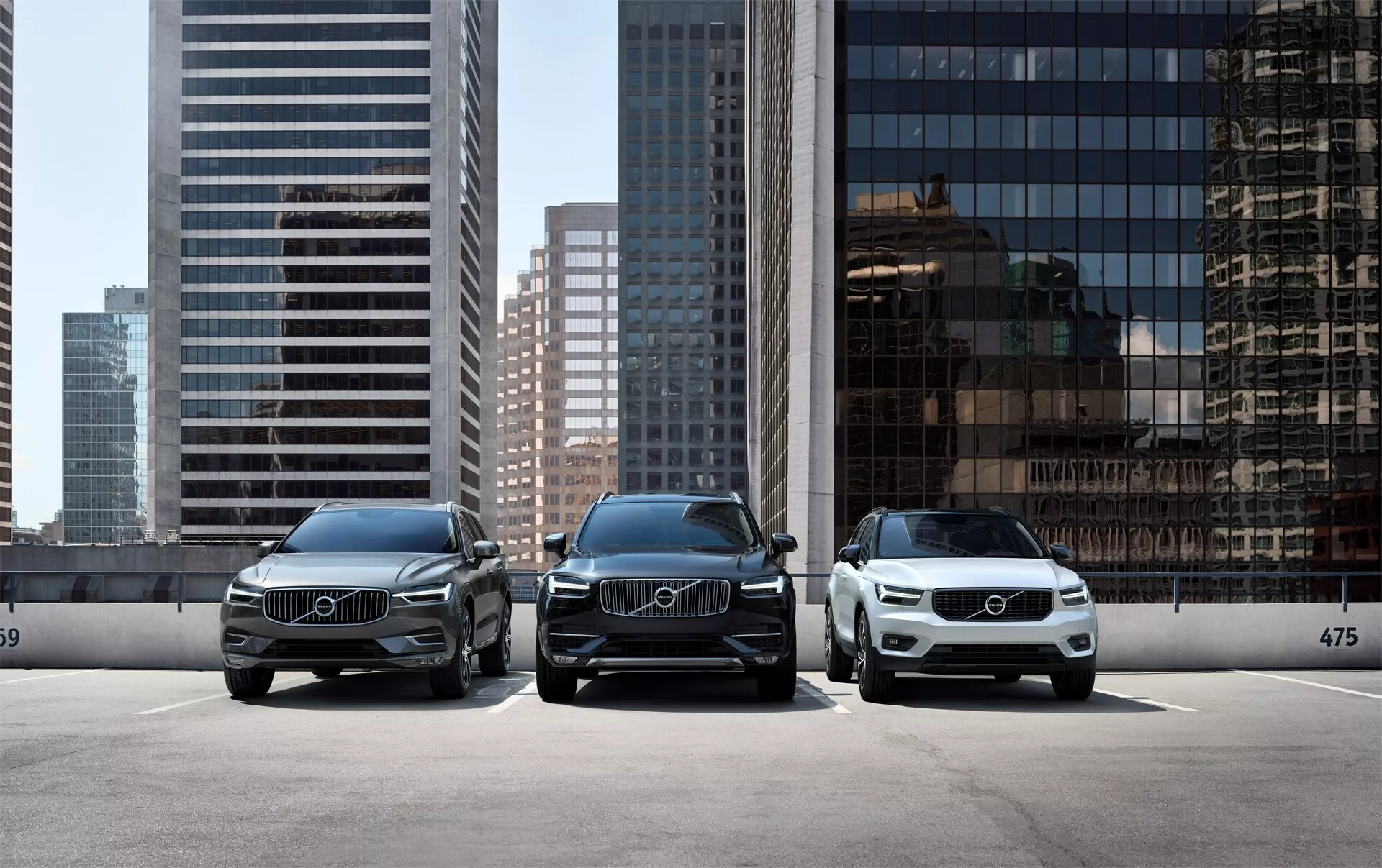 Tri luksuzna avtomobila Volvo XC90 2015 (2. generacija), parkirana vzporedno, obrnjena proti kameri, z modernim mestnim obzorjem v ozadju. Sprednja perspektiva vozil prikazuje elegantne LED luči, velike rešetke z vidnimi Volvo emblemi in čist, eleganten dizajn. Avto v ospredju na desni strani je svetlejše barve, kar poudarja različne barvne možnosti, ki so na voljo pri proizvajalcu. Parkirani so za robnikom s številko 475, kar nakazuje določeno parkirno območje ali razstavni prostor v salonu.