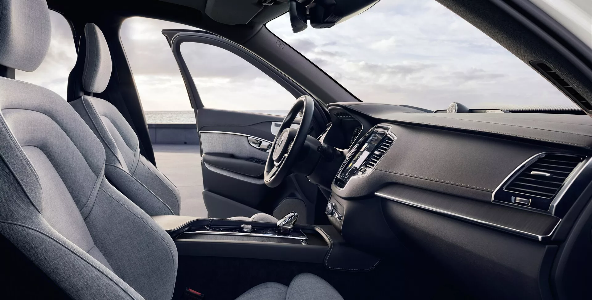 Ta slika prikazuje notranjost druge generacije Volvo XC90 iz leta 2015 s perspektive voznika. Rahlo povišan kot ponuja podroben pogled na sprednja sedeža in armaturno ploščo, s voznikovim sedežem na levi strani, oblazinjenim v teksturirani tkanini z vidnim naslonjalom za glavo in stransko oporo. Večfunkcijski volan ima več kontrolnih gumbov, in elegantna armaturna plošča vključuje simetrične prezračevalne odprtine in delno viden osrednji zaslon za informacije in zabavo. Sredinska konzola prikazuje prestavno ročico za samodejni menjalnik in druge kontrolne funkcije, kar poudarja ergonomsko in praktično zasnovo.