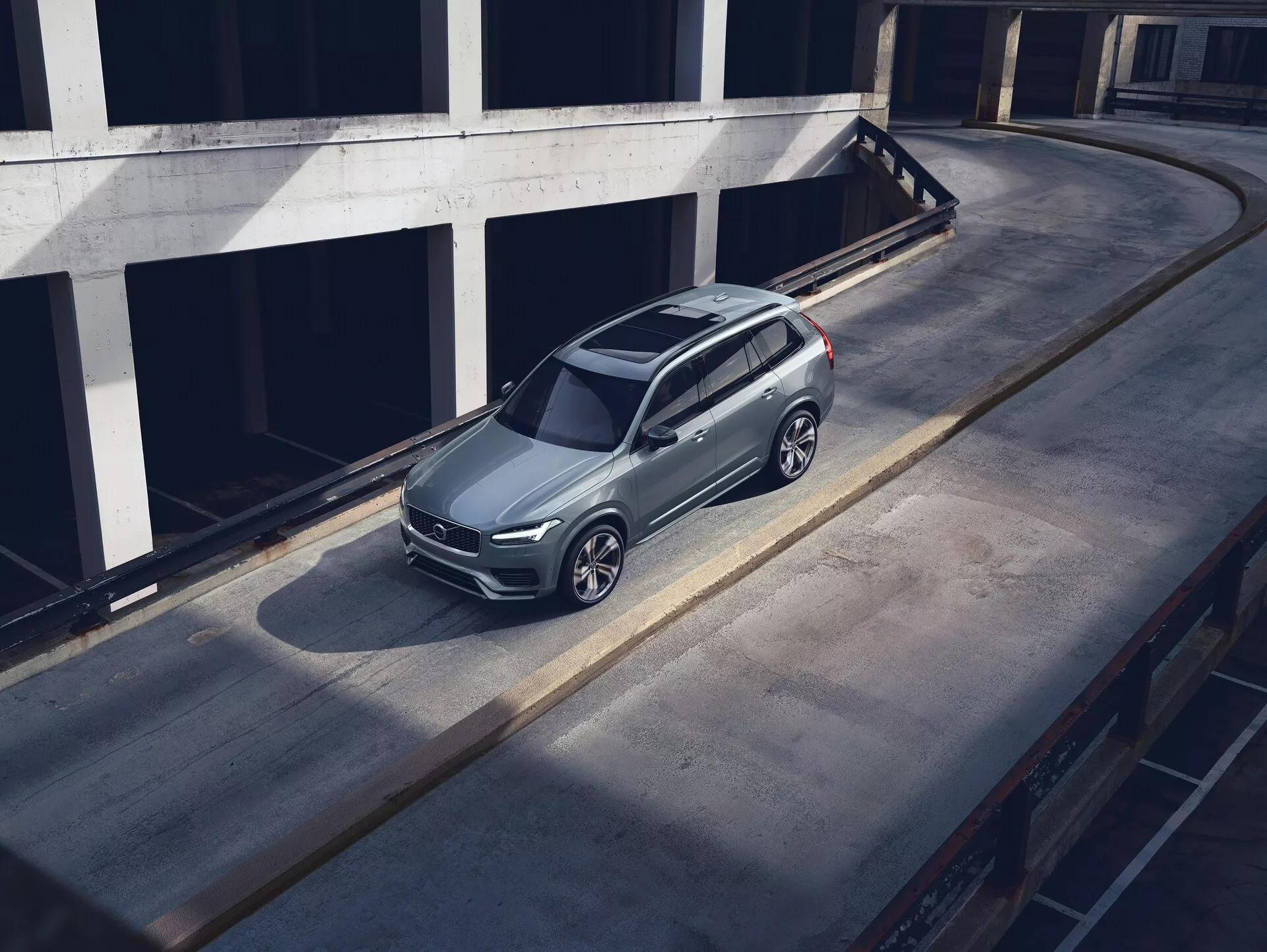 Visok kot ptičje perspektive parkiranega SUV Volvo XC90 iz leta 2015 na betonski površini, ki prikazuje njegov eleganten, sodoben dizajn s kovinskim lakiranjem, značilno sprednjo rešetko, vidnimi žarometi in zapletenimi aluminijastimi platišči. Posnetek poudarja streho, pokrov motorja in levo stran avtomobila, s senco, ki nakazuje jutranjo ali pozno popoldansko svetlobo. Okolje je videti kot zgornja raven večnadstropne garažne strukture, kar povečuje izpostavljenost vozila v čistem, urbanem okolju.