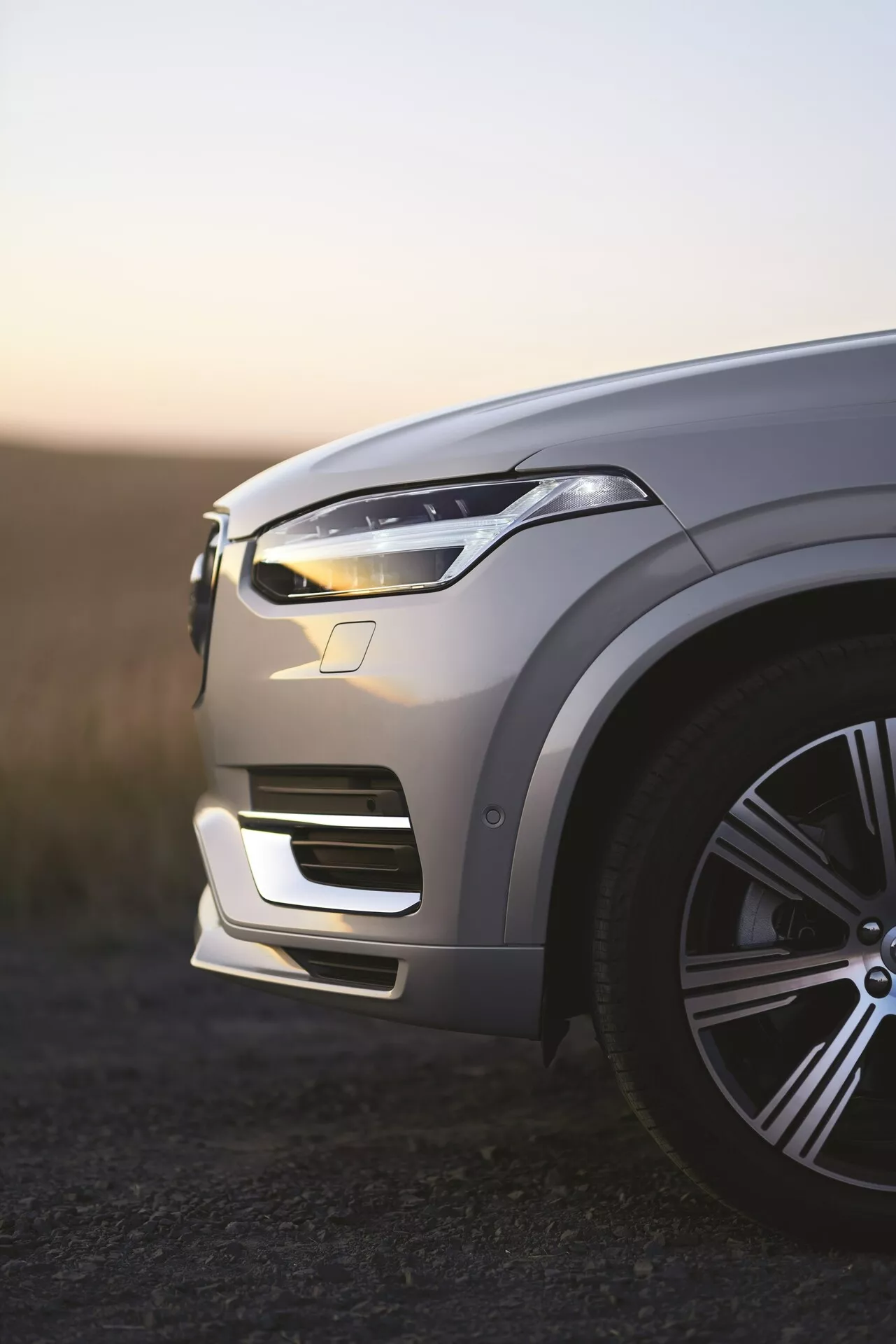 Pogled na levo sprednjo četrtino luksuznega SUV-ja Volvo XC90 druge generacije iz leta 2015 ob zori, osvetljene LED dnevne luči, kovinska barva, ki odseva svetlobo okolice, elegantno veliko lito platišče, zapletene konture pokrova motorja, eleganten dizajn LED žarometa, integriran sprednji odbijač z zračnimi odprtinami, vidni potencialni senzorji za pomoč vozniku, plitka globinska ostrina z zamegljenim ozadjem.
