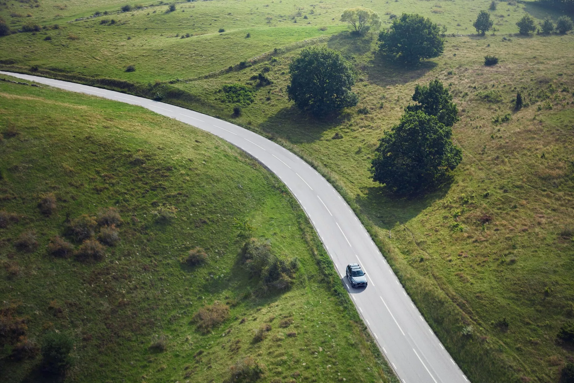 Zračni pogled na srebrni ali svetlo sivi Volvo XC90 iz leta 2015, ki se vozi po podeželski ovinkasti cesti, obdani z zelenimi travnatimi polji z majhnimi drevesi in grmičevjem. Avto, zajet iz ptičje perspektive, je v zavojih na desni strani središča slike, kjer meče senco, kar kaže na opoldansko sonce.