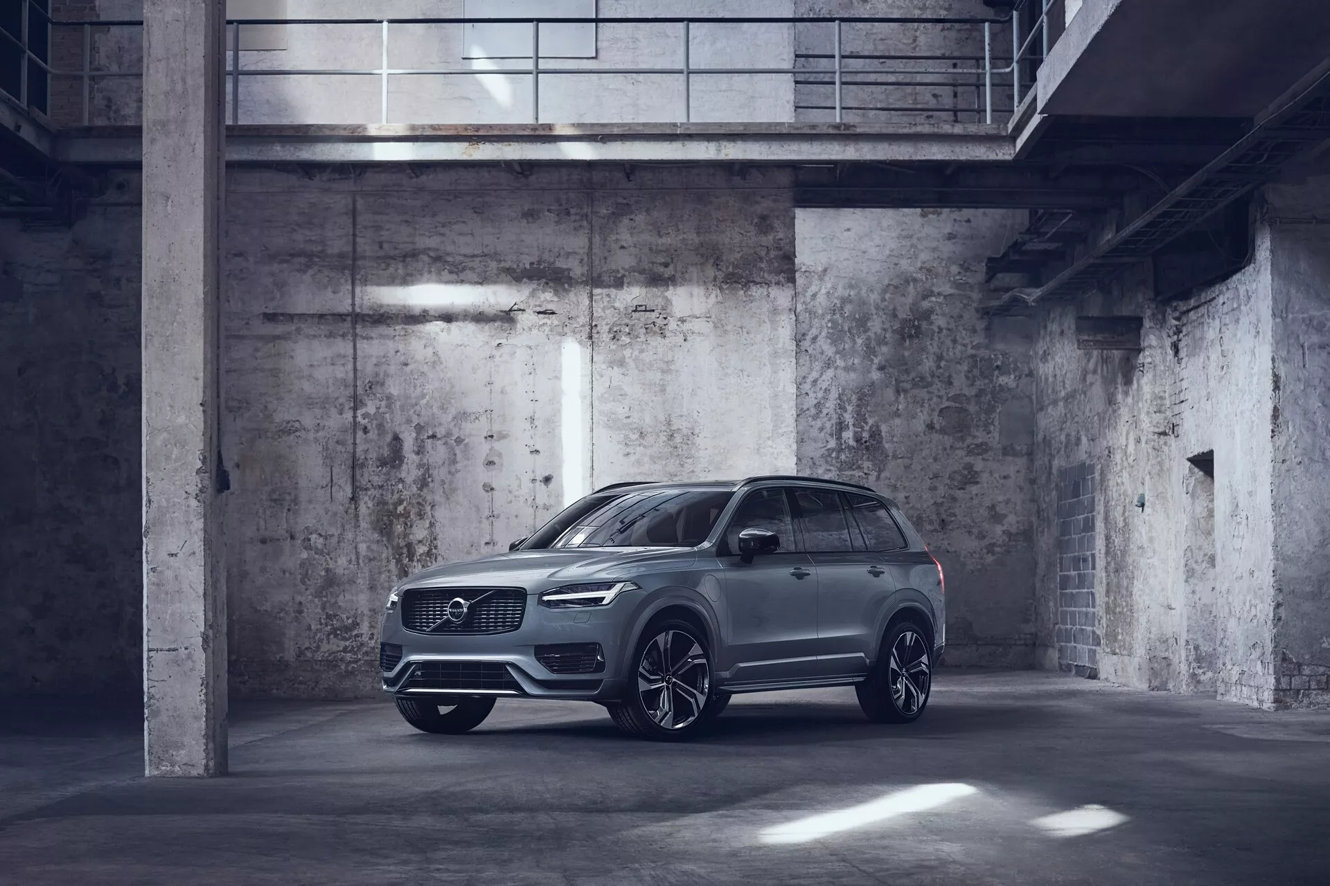Slika kovinsko obarvanega vozila Volvo XC90 2015, parkiranega v slabo osvetljeni, industrijsko oblikovani garaži. Posneta iz tričetrtinskega sprednjega pogleda, prikazuje voznikovo stran z izrazito prikazano spredno stranjo in bočnim profilom. Vozilo ima elegantne LED žaromete, veliko sprednjo mrežo, verjetno parkirne senzorje in velika lita platišča z večkrakim dizajnom. Stranska ogledala so aerodinamične oblike in lahko vključujejo napredne tehnološke funkcije, kot so kazalniki smeri in sistemi za opozarjanje na mrtvi kot. Slika poudarja luksuzno, a praktično zasnovo SUV-ja z visoko oddaljenostjo od tal in možnimi stropnimi funkcijami, kot je strešno okno ali panoramska streha.