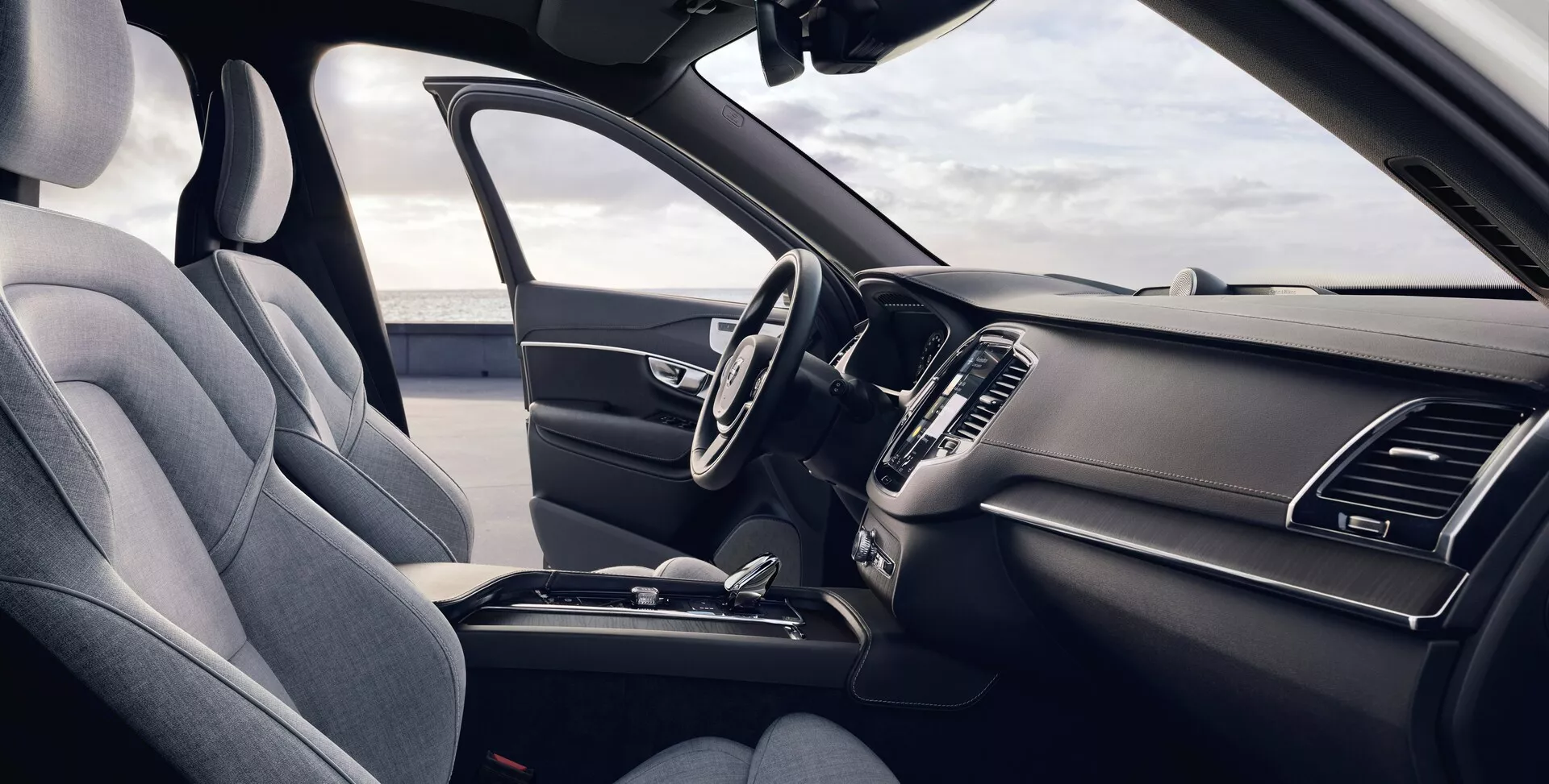 Pogled na notranjost Volvo XC90 2015 (2. generacija) iz perspektive sprednjega sopotnika prikazuje ergonomski sedež voznika, oblazinjen s temnim premium blagom ali sintetičnim usnjem, večfunkcijskim volanom z nameščenimi kontrolami, eleganten armaturni plošči z izključenim infotainment sistemom na dotik, vgrajenimi zračniki, minimalističnimi klimatskimi kontrolami, osrednjo konzolo s shranjevalnim predalom, držali za pijačo in prestavno ročico, vse izpostavljeno v enotnem temnem barvnem vzorcu z naravno dnevno svetlobo, ki osvetljuje vozilo.