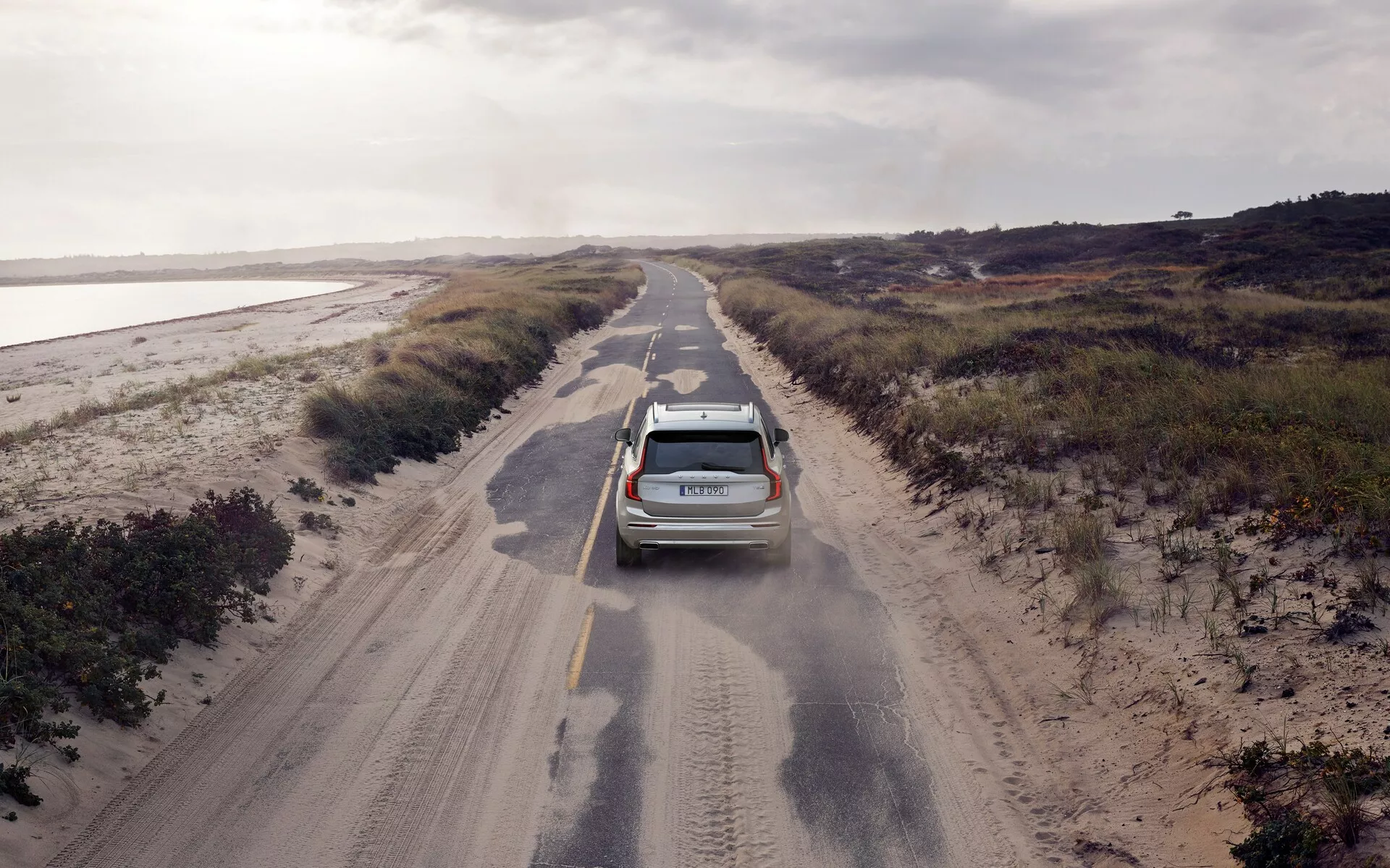 Pogled iz zraka na SUV Volvo XC90 2015 (2. generacija), ki vozi po ozki, neasfaltirani cesti, obdani z nizko vegetacijo. Sodobno vozilo, opremljeno z anteno in strešnimi letvami, je nekoliko zamaknjeno v levo in dviguje prah, kar kaže na gibanje in grobo teksturo ceste. Oblačna osvetlitev enakomerno osvetli prizor, kar prispeva k občutku izolacije. Vidne značilnosti vključujejo zadnje luči, zadnji brisalec in morda parkirne senzorje.