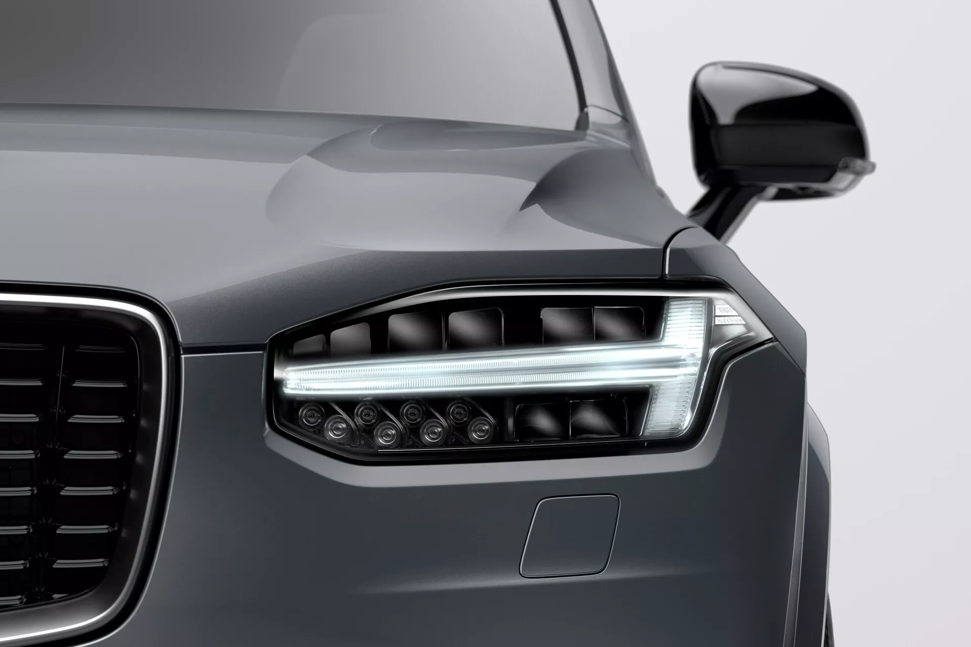 Pogled od blizu na levi sprednji kot Volva XC90 2015 (2. generacija), prikazano moderno LED zasnovo žarometov z vogalnimi dnevnimi lučmi, delni pogled na veliko rešetko z navpičnim vzorcem, sijajna temno siva kovinska barva in parkirni senzor na odbijaču, posneto iz nizkega kota od desne proti levi.