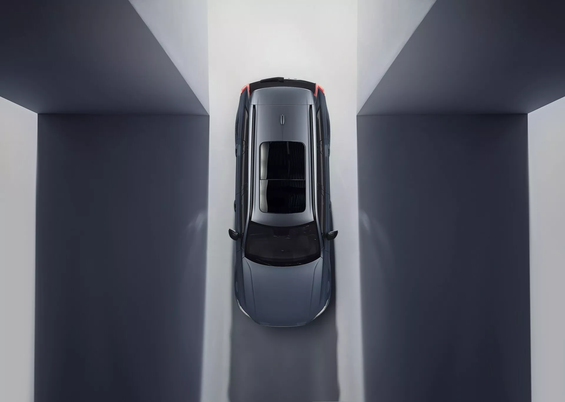 Pogled iz zraka na Volvo XC90 2015 (2. generacije), ki prikazuje simetrično kompozicijo in odseve okolja. Osrednja postavitev v notranjem prostoru z nevtralnimi toni poudarja streho avtomobila s strešnim ali panoramskim steklom. Vidni deli vključujejo pokrov motorja, streho, prtljažnik in zgornjo polovico vrat, kar poudarja elegantne linije in krivulje, ki nakazujejo luksuzno zasnovo, osredotočeno na aerodinamičnost in prostorno notranjo osvetlitev.