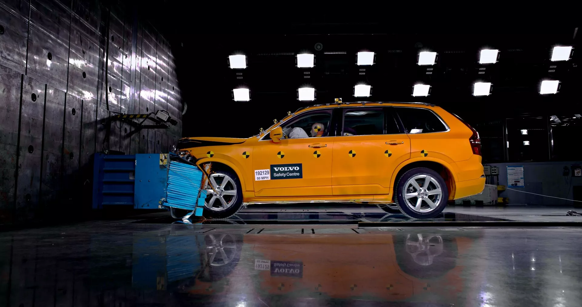 Bočni profil svetlo rumene Volvove XC90 iz leta 2015 (2. generacija) v industrijskem okolju, ki prikazuje njegov karavanski dizajn z vidno sprednjo mrežo, žarometi in aluminijastimi platišči. Avto je povezan z modro diagnostično ali polnilno napravo, lutka znotraj pa nakazuje varnostno ali trkalno testiranje. Polirana tla odsevajo podvozje in kolesa na nasprotni strani.
