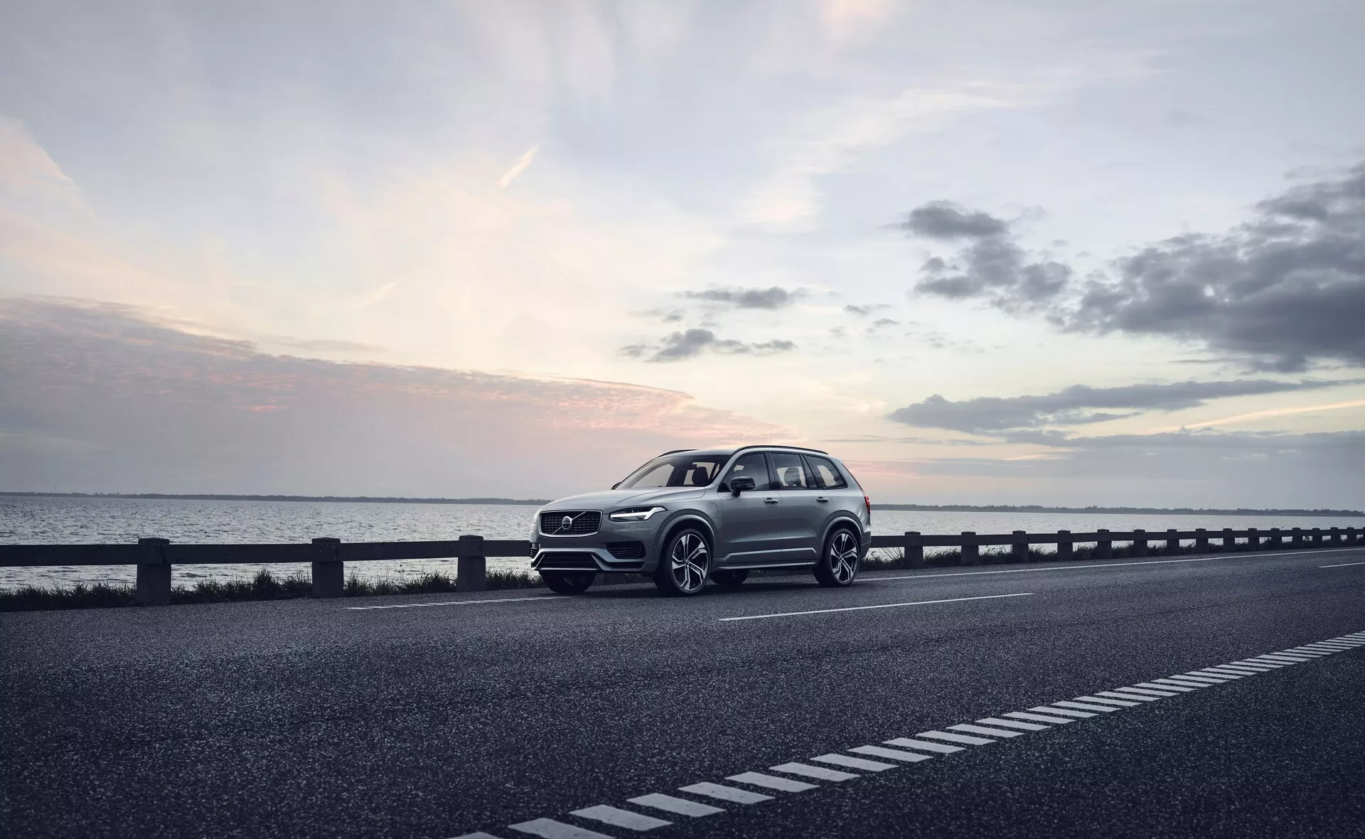 Sodobni SUV, natančneje model Volvo XC90 iz leta 2015 (2. generacija), parkiran na desni strani dvosmerne ceste. Vozilo je fotografirano iz tridčetrtinske sprednje perspektive, ki prikazuje sprednjo masko, žaromete, pokrov motorja in prednji blatnik. Kot razkriva tudi del profila avtomobila, vključno s kolesi, voznikovimi vrati in delom zadnjega dela. Kolesa so rahlo obrnjena v desno, z morebitnimi športnimi oblikovnimi elementi, kot je izbočen odbijač in večji zračniki, kar nakazuje na dinamični paket. Fotografija, verjetno posneta ob mraku z mehko, razpršeno svetlobo pod rahlo oblačnim nebom, povečuje mirno vzdušje. Dobro definirana karoserija z vidnimi značilnimi črtami nakazuje na prestižni model. Tehnološke značilnosti, ki jih lahko razberemo iz slike, vključujejo LED žaromete, napredne dizajne koles, prefinjeno sprednjo masko z morebitnimi senzorji ali kamerami ter aerodinamične podrobnosti. Ta visokokakovostna slika bi bila primerna za oglaševanje, poudarjajoč avto kot luksuzno izbiro življenjskega sloga.