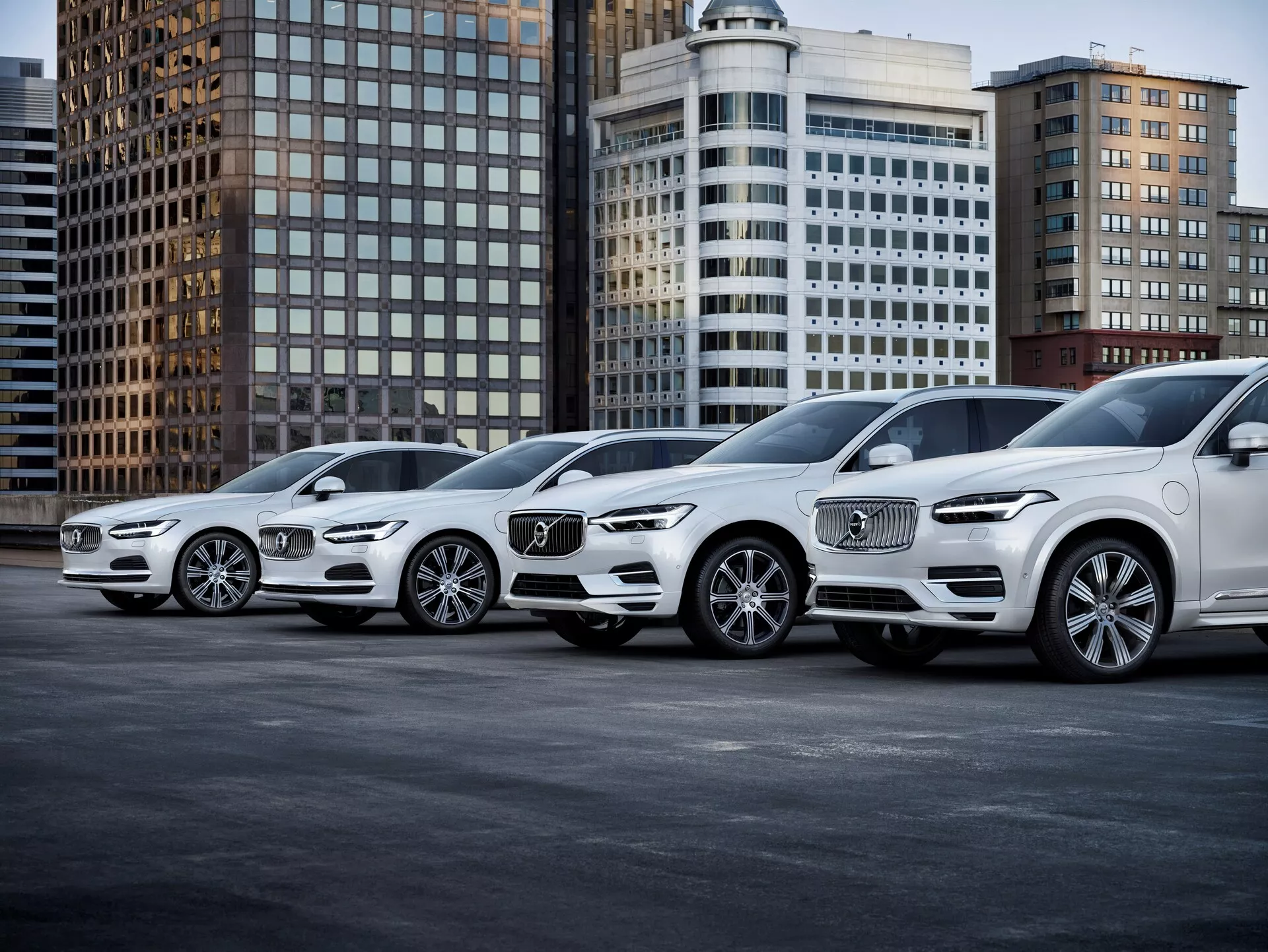 Slika prikazuje vrsto štirih luksuznih SUV-jev Volvo XC90 druge generacije, letnik 2015, s sprednjimi profili usmerjenimi v desno. Vozila so postavljena diagonalno, perspektiva pa poudarja tako sprednje kot stranske poglede vozil. Avtomobili imajo izrazite sprednje rešetke s prepoznavnim Volvovim logotipom, zapletene LED žaromete, oblikovanje zlitinskih koles in obrisne linije na pokrovu motorja. Fotografija, posneta iz nizkega kota blizu tal, poudarja velikost in držo SUV-jev, postavljenih na urbano ozadje z pisarniškimi stavbami. Osvetlitev je mehka, kar nakazuje oblačen dan ali dobro osvetljeno urbano okolje, jasno prikazuje kolesa, žaromete, meglenke in oblikovalske črte. Najbližje vozilo prikazuje napredne osvetlitvene tehnologije in prednje senzorje na rešetki, verjetno povezane z avtonomno vožnjo ali parkirno asistenco. Ko se avtomobili umaknejo, njihov enoten stil kaže, da pripadajo isti družini vozil, zaradi česar je ta slika idealna za kategorije, kot so Avtomobilizem, Luksuzna vozila, Napredne tehnologije vozil in Mestni življenjski slog.