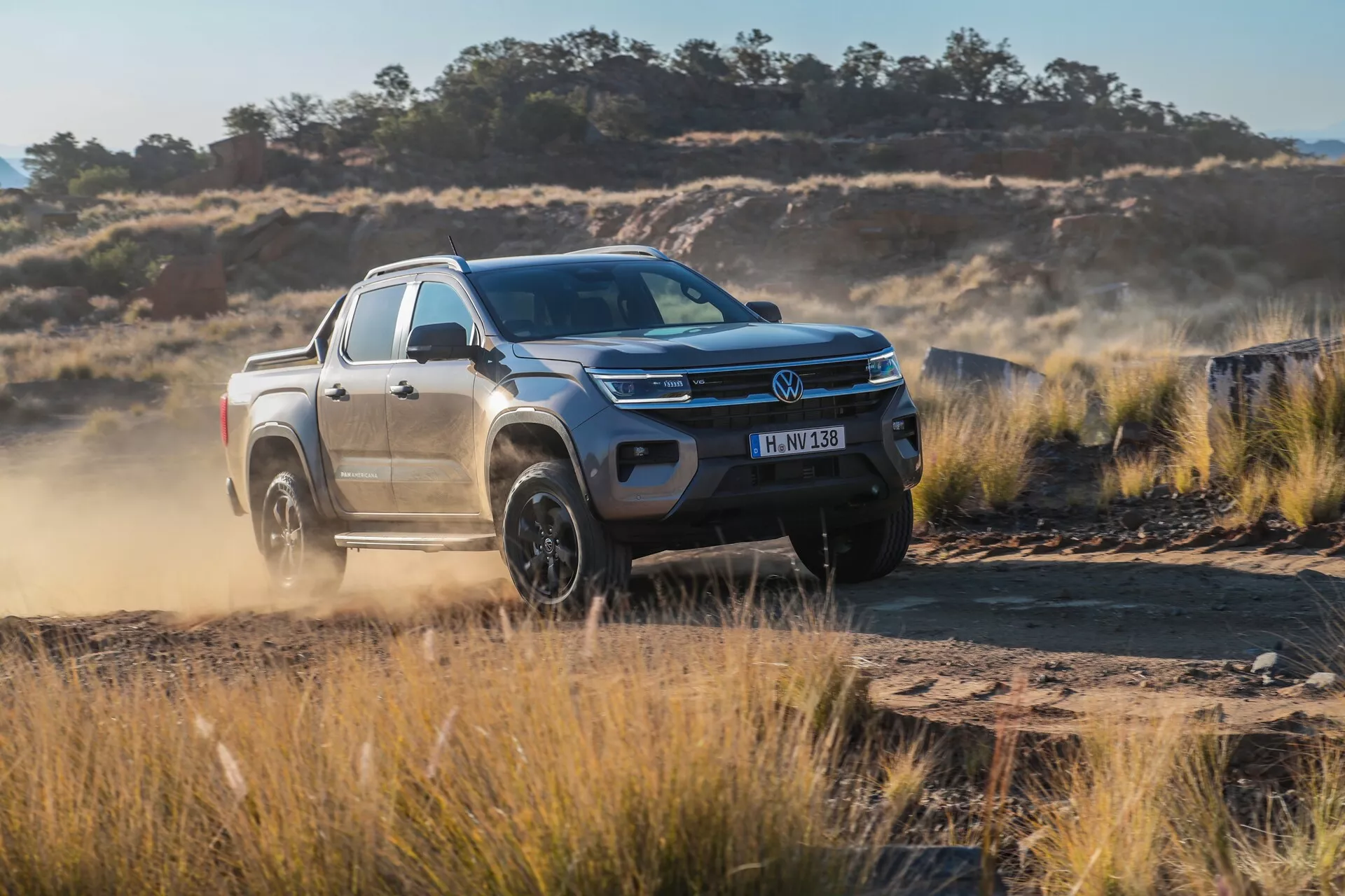 Trikrat spredaj gledan pogled na pickup Volkswagen Nutzfahrzeuge Amarok 2023 (2. generacija), ki prikazuje njegov dinamičen dizajn na robustni makadamski cesti. Slika prikazuje LED žaromete tovornjaka, s kromom zaključeno rešetko z logotipom proizvajalca, oblikovano pokrov motorja, robusten sprednji odbijač, velika platišča iz zlitine in dodatke, kot so stranska ogledala z vgrajenimi smerniki, strešne sani in prečnik. Tovornjak je prikazan v gibanju, kar poudarja njegove terenske zmogljivosti s prahom, ki se dviga za njim, kar nakazuje napredne sisteme za nadzor oprijema in zmogljivosti pogona na štiri kolesa.