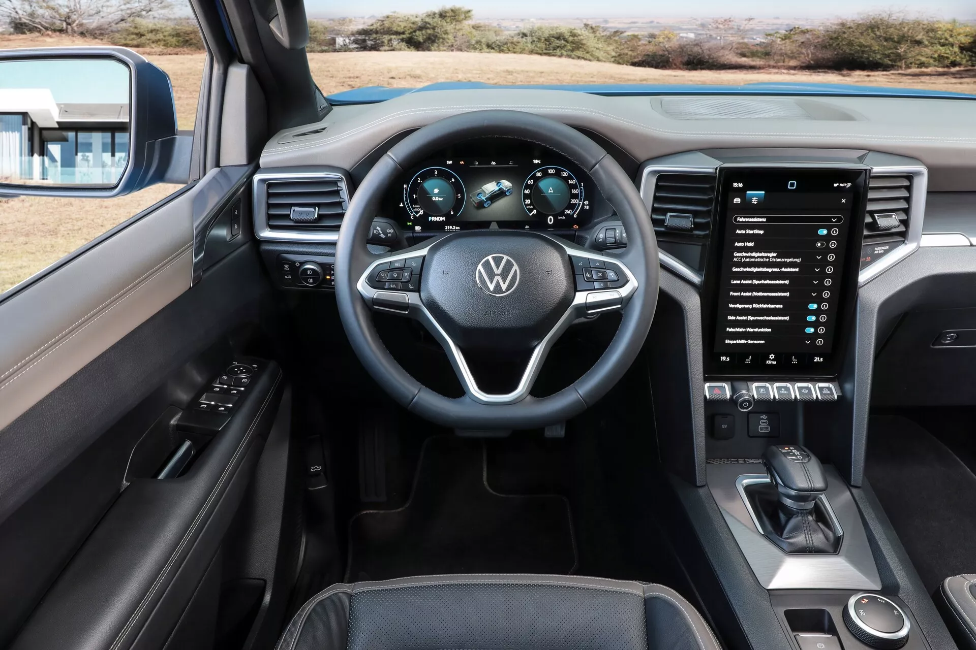 Pogled z notranjosti z voznikovega sedeža na armaturno ploščo, volan, instrumentno ploščo in informacijski sistem vozila Volkswagen Amarok 2023 (2. generacija). Večfunkcijski volan z logotipom VW in kontrolnimi gumbi je na sredini, zadaj pa je digitalna instrumentna plošča, ki prikazuje hitrost in vrtljaje. Velik vertikalni informacijski zaslon na dotik prevladuje nad osrednjo konzolo, obkrožen je z gumbi za upravljanje klime in sodobno prestavno ročico, prikazuje eleganten in prefinjen dizajn armaturne plošče s premium materiali.