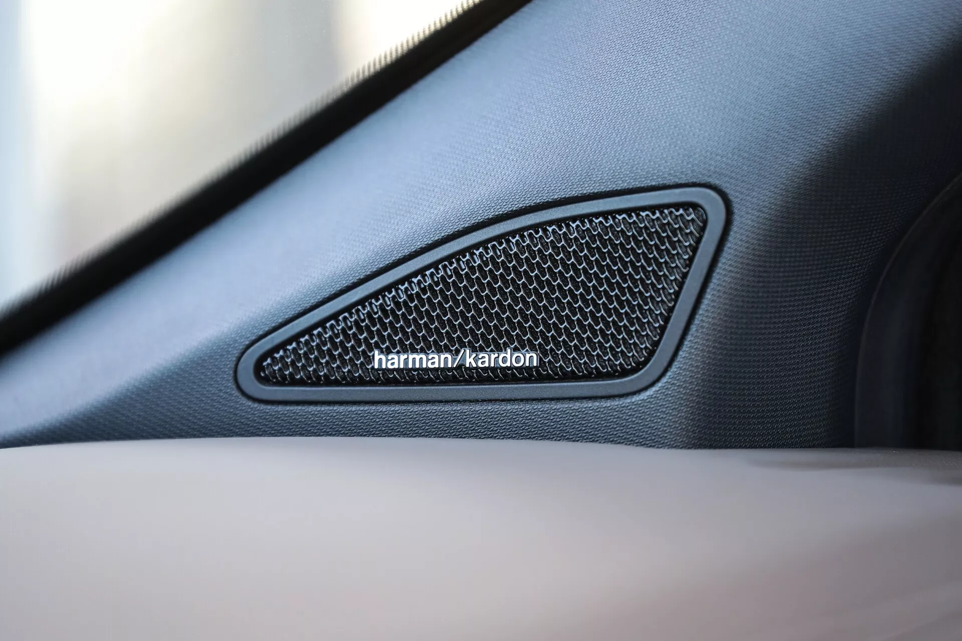 Povečava mreže notranjega zvočnika VW Nutzfahrzeuge Amarok 2023 na vratnem panelu; trikotna zasnova se zoži proti sprednjemu delu, harman/kardon znamčenje, šesterokotna mrežasta tekstura in finozrnata usnjena obloga; poudarek na vrhunskem avdio sistemu z majhno globino ostrine, ki poudarja podrobnosti zvočnika.