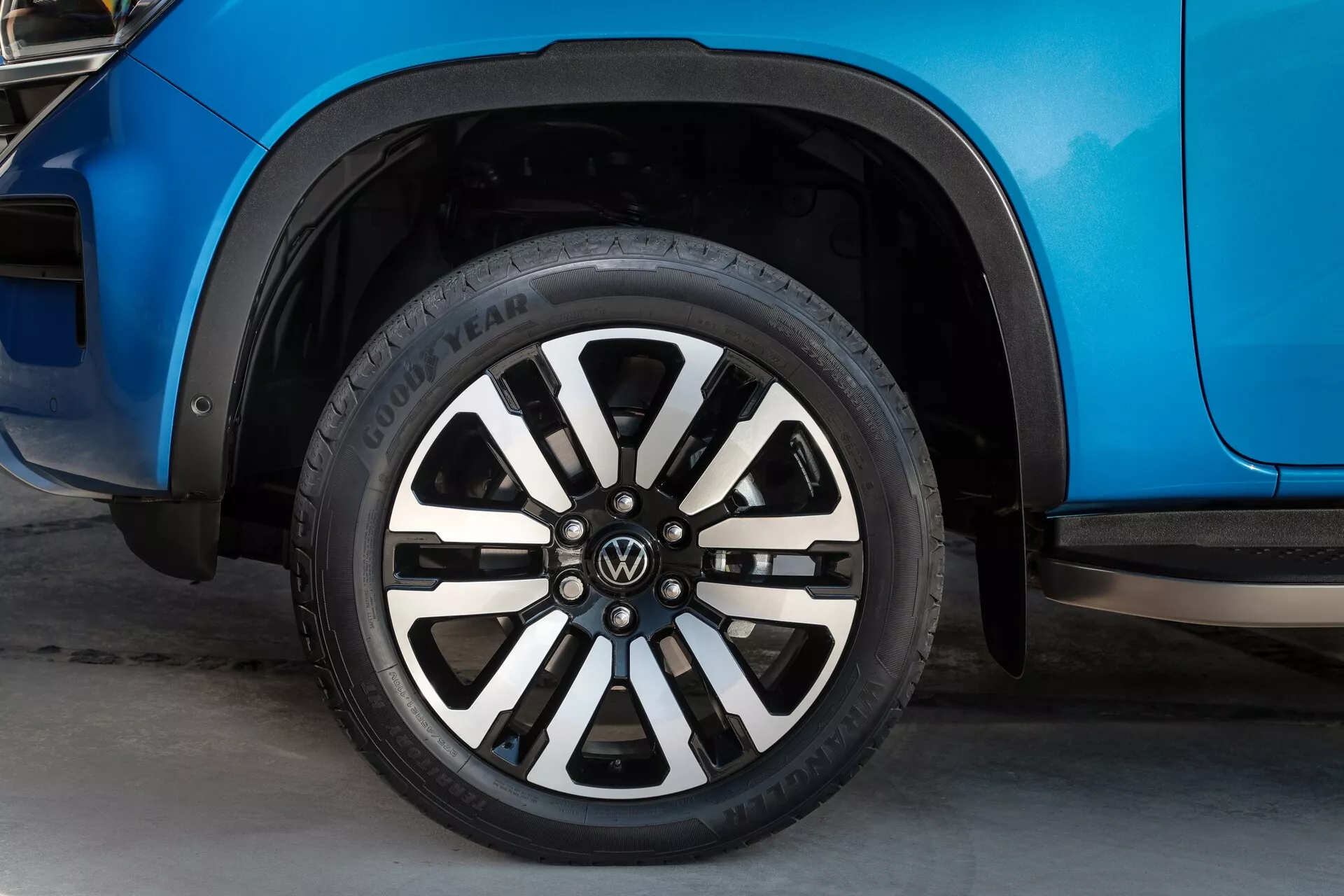 Posnetek od blizu, osredotočen na kolo druge generacije VW Nutzfahrzeuge Amarok 2023. Slika prikazuje desno sprednjo stran vozila, vključno z delom kolesnega loka, sprednjega vratnega panela in črnih plastičnih zaščitnih blatnikov. Kolo ima večkraki aluminijasti dizajn z izmenjujočimi se črnimi in srebrnimi kraki ter logotipom VW na sredini. Vidna je tekalna plast pnevmatike Goodyear, karoserija vozila pa ima živahno modro kovinsko barvo, ki odbija svetlobo. Posnetek je narejen iz nizkega kota, z rahlo zamegljenim ozadjem, kar poudari pnevmatiko in platišče.