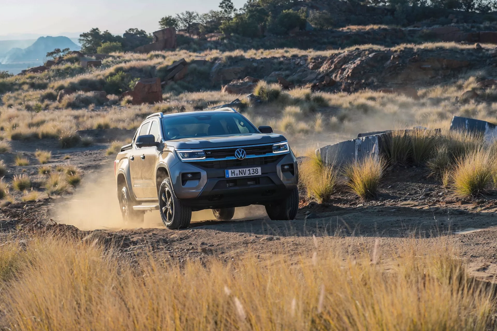 Volkswagen Amarok 2023 (2. generacija) pickup prikazan iz sprednjega trikotnega pogleda v terenskem okolju. Vozilo prikazuje sodobne oblikovalske elemente, kot so integrirane LED dnevne luči in močan sprednji odbijač. Fotografija poudarja višjo oddaljenost Amaroka od tal in zasnovo aluminijastih platišč, kar poudarja njegovo primernost za zahtevne razmere. Vidni prah, ki ga dvigujejo zadnja kolesa, prikazuje njegove terenske zmogljivosti, zaradi česar je idealen za pustolovce in tiste, ki iščejo uporabnost in slog.
