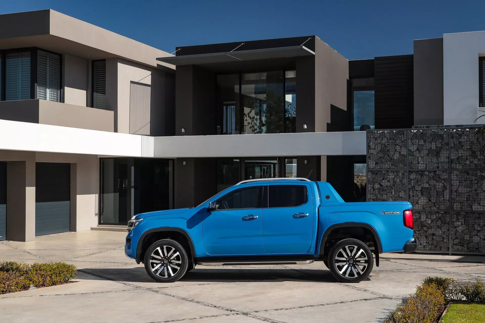 Slika 2023 VW Nutzfahrzeuge Amarok druge generacije, modernega modrega poltovornjaka s kovinsko barvo, parkiranega na dovozu stanovanjske hiše, s prefinjenim predmestnim ozadjem. Položaj iz tričetrtinskega kota prikazuje profil tovornjaka, vključno z obliko kabine, kesona in oblokov koles, z namigi na zasnovo sprednje maske in žarometov. Velika lita platišča nakazujejo kombinacijo uporabnosti in razkošja. Fotografija je posneta na srednji razdalji pri naravni dnevni svetlobi, kar poudarja poliran videz vozila in oblikovne značilnosti, kot so LED žarometi, tehnologije za pomoč vozniku in konfiguracija kabine za ekipo s štirimi vrati.