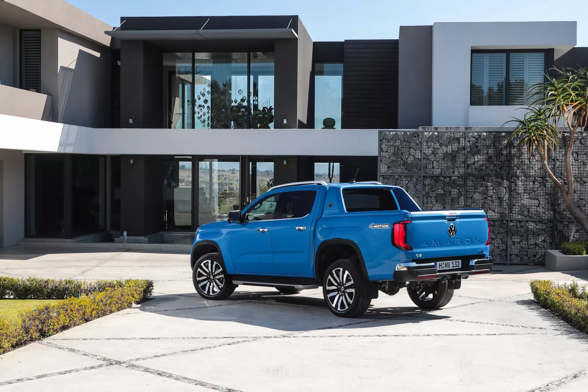 Trikratni zadnji pogled na sodoben modri pick-up VW Nutzfahrzeuge Amarok 2023, ki prikazuje svoja lita kolesa, sijočo kovinsko barvo, zadnje senzorje, LED zadnje luči in anteno za plavut morskega psa. Vozilo je parkirano na tlakovanem dovozu pred sodobno luksuzno rezidenco, kar poudarja njegovo uporabnost v vrhunskem stanovanjskem okolju.