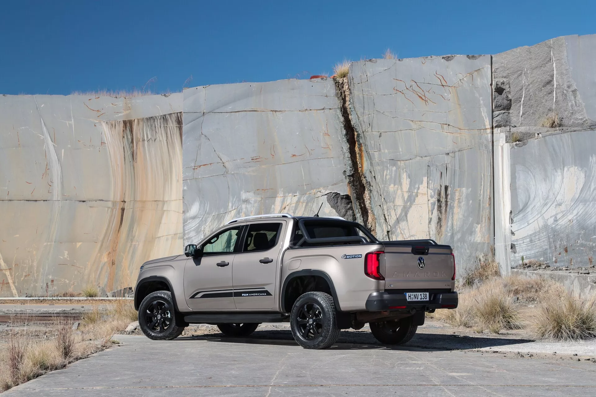 Trikrat četrti zadnji pogled na pickup VW Nutzfahrzeuge Amarok 2023 (2. generacija), ki prikazuje njegove stranske in zadnje oblikovne elemente. Tovornjak je postavljen na ravno betonsko površino proti grobi, teksturirani kulisi. Ima mat sivo ali bež barvo z črnimi poudarki na kolesih, ogledalih in okvirjih, kar predlaga posebno izdajo ali off-road paket. Funkcije vključujejo aluminijasta kolesa, terenske pnevmatike, konfiguracijo z dvojno kabino s štirimi vrati in zadnja vrata z znak proizvajalca in vlečnim priklopom. LED zadnje luči in pokrov kesona prispevajo k njegovemu sodobnemu, funkcionalno usmerjenemu dizajnu.
