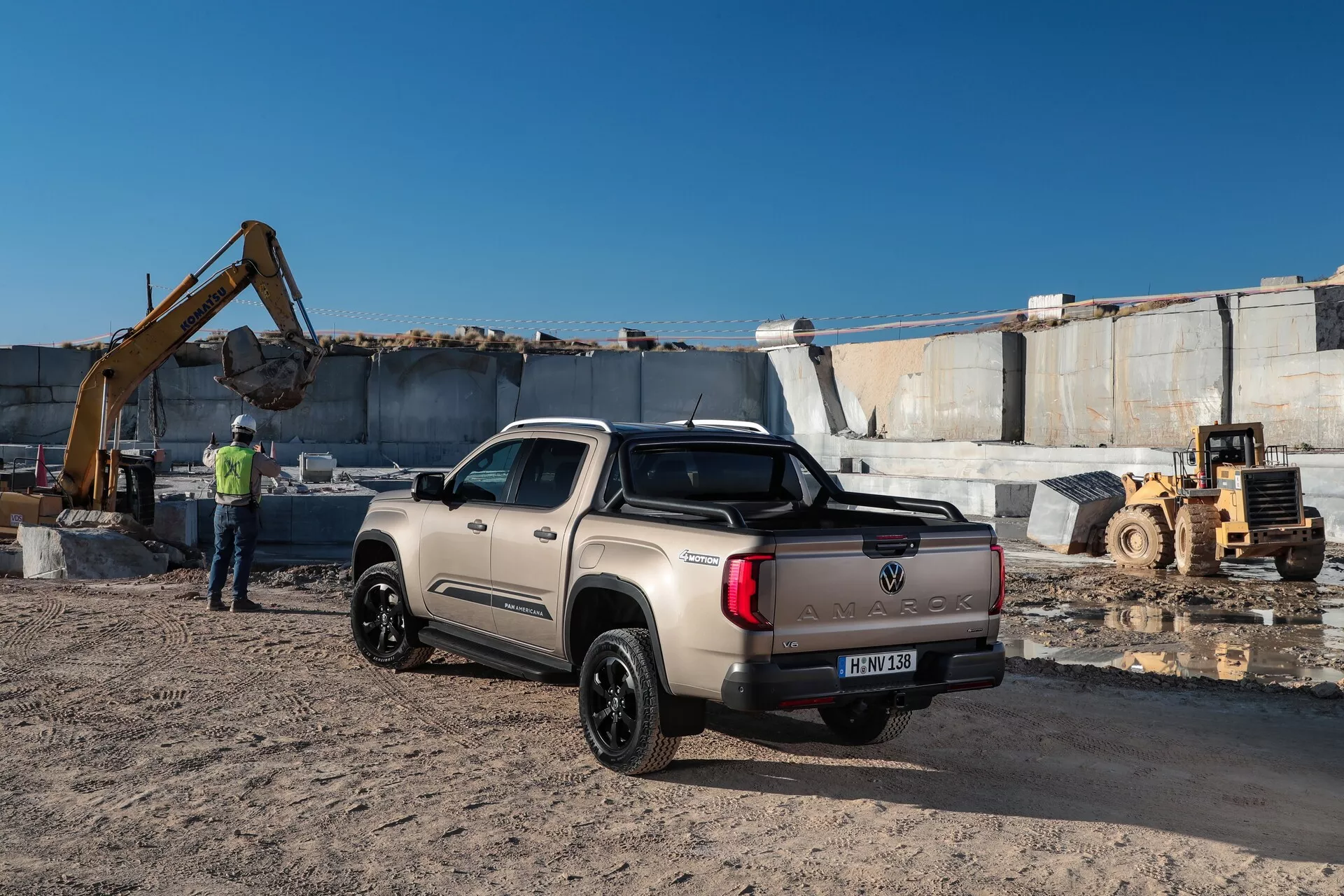 Slika pickup tovornjaka VW Nutzfahrzeuge Amarok 2023 v industrijskem okolju z izkopno opremo in velikimi kamnitimi bloki v ozadju. Posnet z zadnjega trikratrskega kota, svetlo barvani tovornjak s črnimi obrobami in kolesi je viden v celoti z dvojno kabino in pokritim tovornim prostorom. V ozadju je vidna oseba v odsevnem jopiču, kar poudarja gradbiščni kontekst.