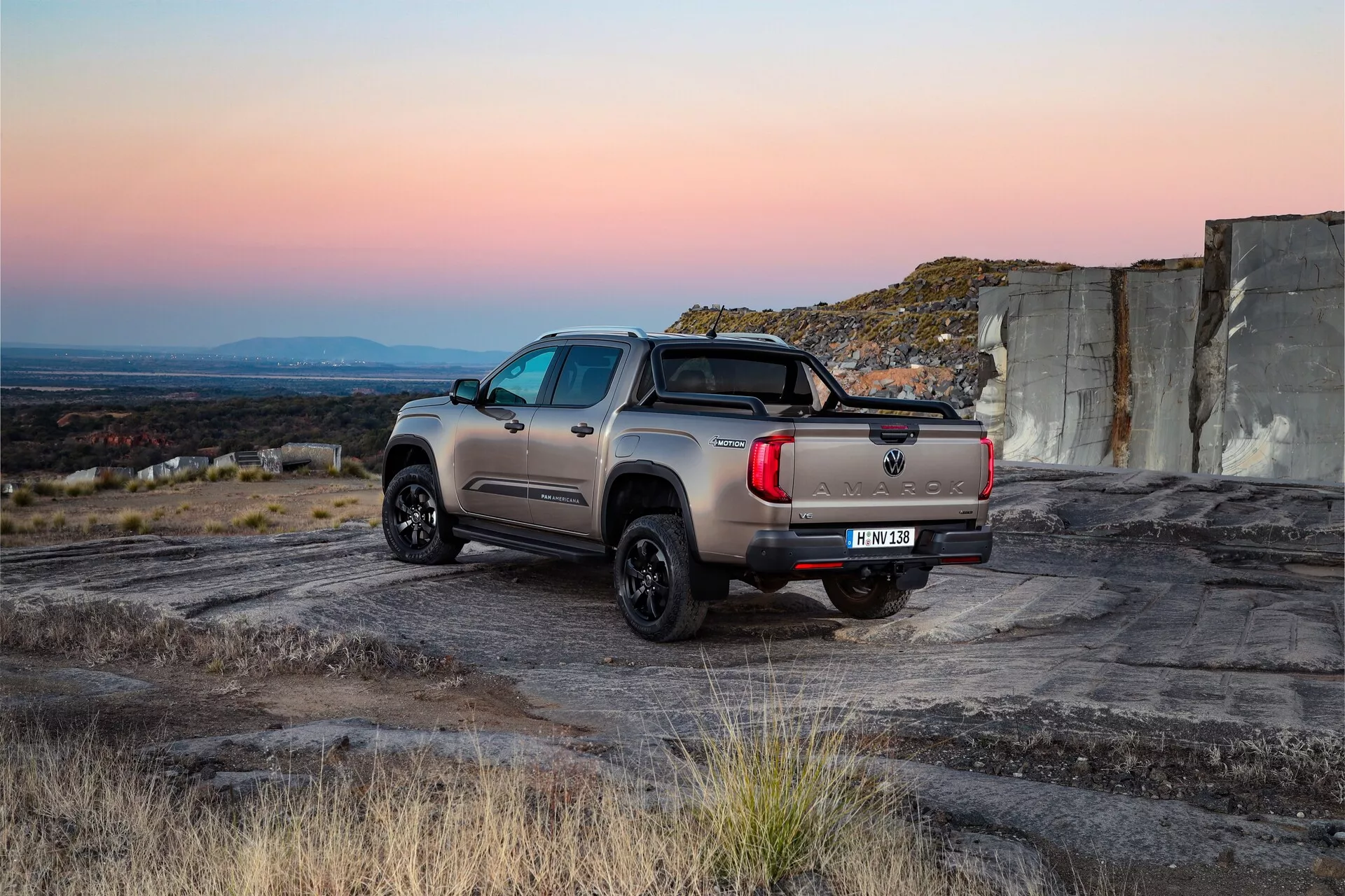 Zadnji trikotni pogled na VW Nutzfahrzeuge Amarok 2023, prikazuje robusten zunanji dizajn na grobem terenu. Sodobni pickup ima močno telo z nemetalno barvo, sodobne LED zadnje luči in vlečno kljuko pod zadnjim odbijačem. Črna, pobarvana aluminijasta platišča s terenskimi pnevmatikami poudarjajo njegovo robusno uporabnost, medtem ko športna palica v postelji tovornjaka dodaja estetski privlak in funkcionalnost za pritrditev tovora. Stranske stopnice in zatemnjena zadnja okna izboljšujejo dostopnost in zasebnost. Položaj vozila v naravni svetlobi s toplimi odtenki in hladnimi sencami poudarja njegove zmogljivosti in uporabniške lastnosti.
