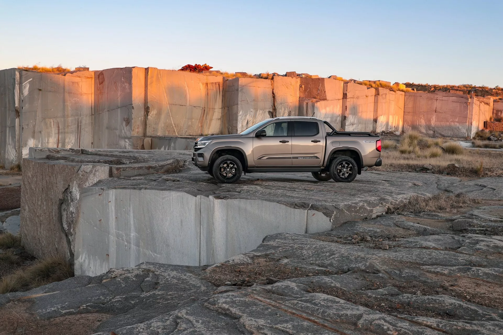 Srednje velik pickup VW Nutzfahrzeuge Amarok 2023 (2. generacija) na skalnatem terenu, verjetno v odprtem kopu. Vozilo je prikazano iz zadnjega kota pod kotom treh četrtin v poznih popoldanskih urah. Slika poudarja konfiguracijo dvokabine, svetlo utišano barvo, sodoben dizajn z LED zadnjimi lučmi, oblikovano zadnjo loputo, značilna kolesa in pnevmatike z agresivnim profilom, kar poudarja terenske zmogljivosti.