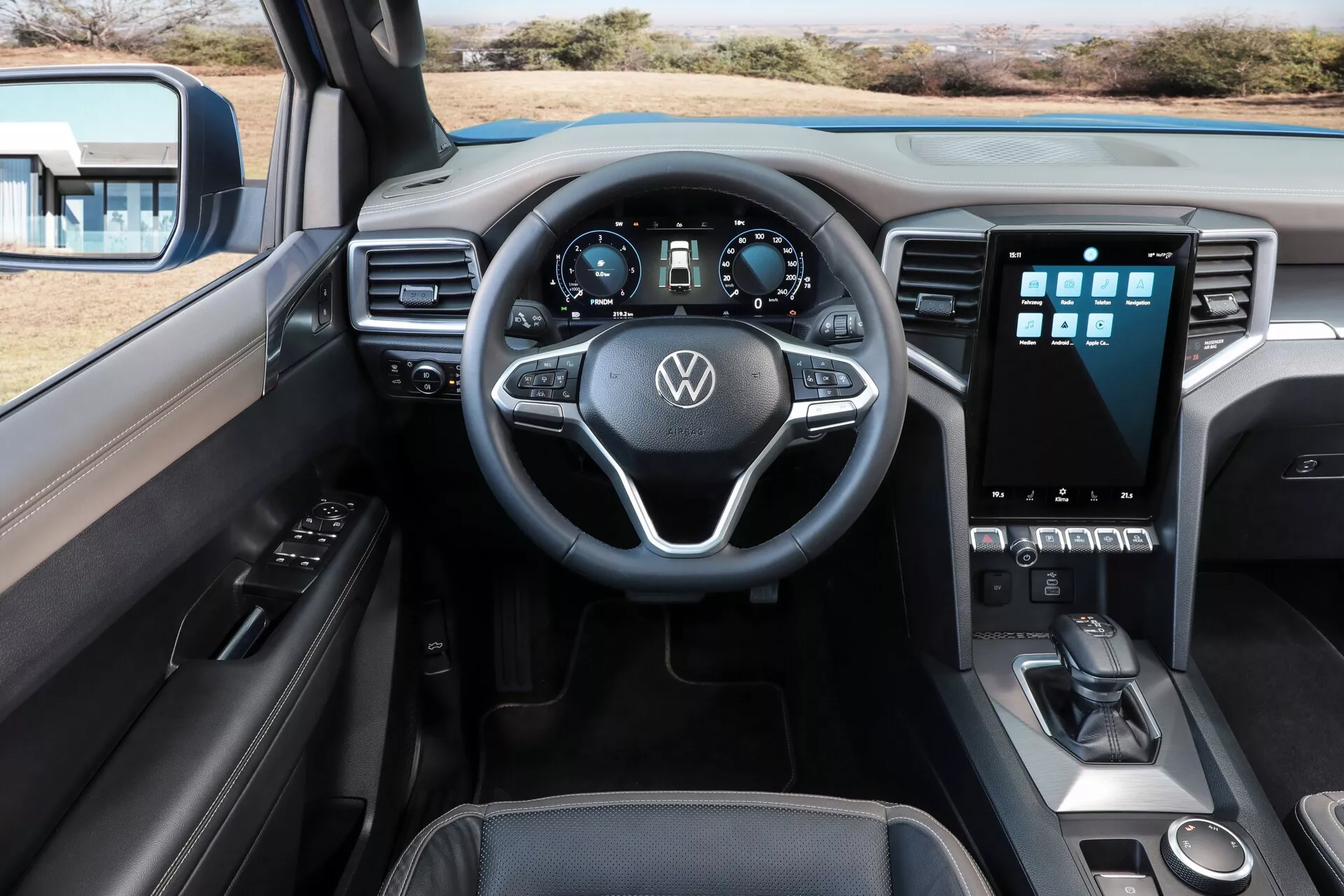 Notranjost Volkswagen Nutzfahrzeuge Amarok 2023 (2. generacija), zajeta iz voznikove perspektive, poudarja armaturno ploščo, volan in sredinsko konzolo. Tri-kraki volan vključuje vgrajene kontrole za avdio, tempomat in druge sisteme, medtem ko digitalni instrumentni panel prikazuje osnovne informacije o vožnji. Sredinska konzola vključuje pokončno usmerjen zaslon na dotik infotainment sistema za navigacijo in nastavitev vozila, obdan s fizičnimi gumbi in gumbi za nadzor klime. Spodaj so vidni upravljalniki za menjalnik in vozne načine ter majhen prostor za shranjevanje. Dizajn izžareva modernost z usnjenimi ali mehkimi materiali in kovinskimi poudarki. Voznikova vrata imajo kontrole za okna in ogledala, s pogledom na usklajeno oblazinjenje na sovoznikovem sedežu.