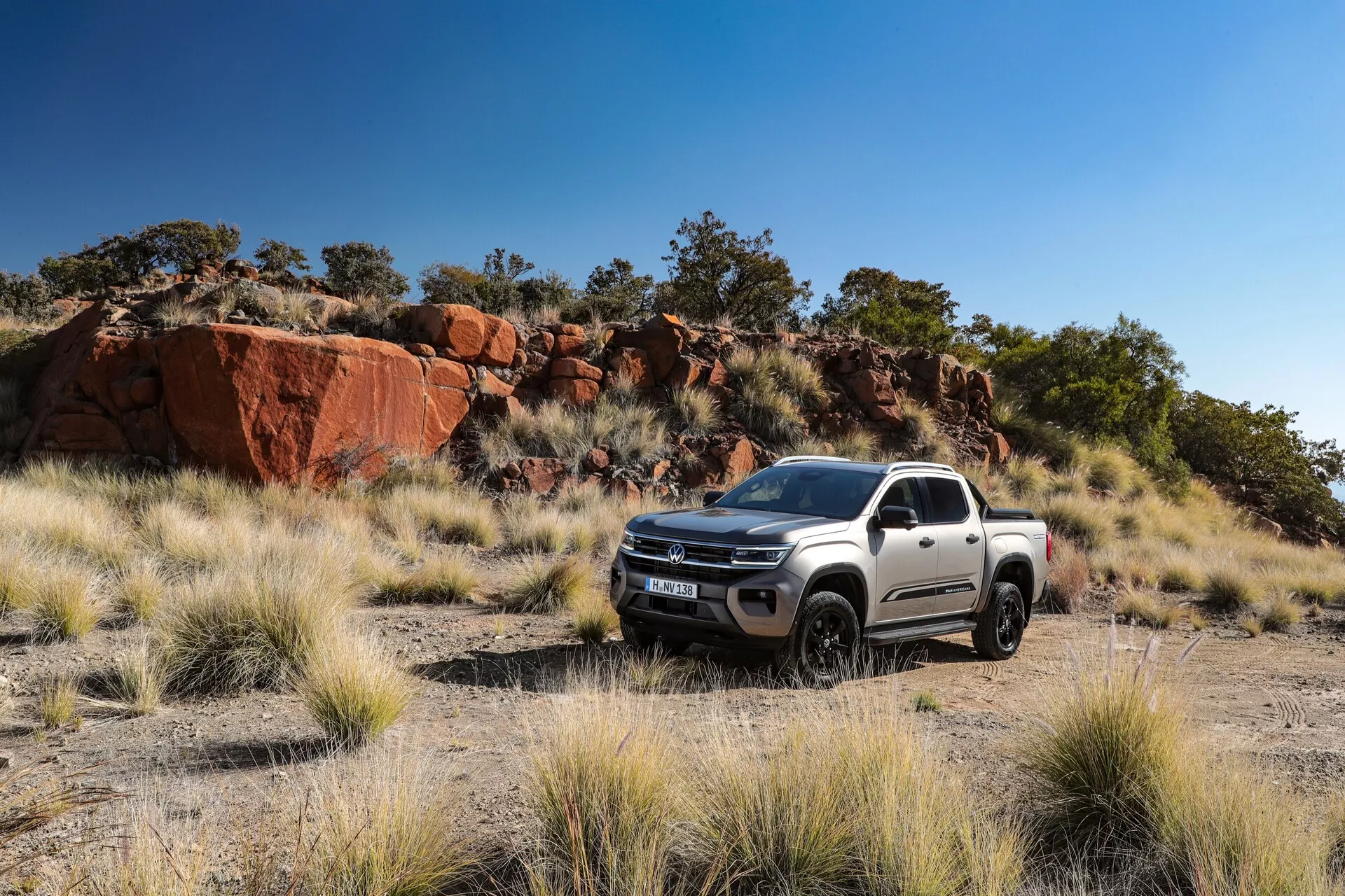 Slika belega pick-upa VW-Nutzfahrzeuge Amarok 2023 druge generacije v naravnem okolju s travnato površino in skalnim terenom, ki poudarja njegove terenske sposobnosti. Fotografirano iz tridelnega prednjega kota prikazuje rešetko, žaromete, sprednji odbijač, pokrov motorja in kolesa na strani voznika. Sodobna zasnova in robustne konture poudarjajo trpežnost, z aluminijastimi platišči, vidnimi sestavnimi deli vzmetenja in zadostno oddaljenostjo od tal. Naravna svetloba na sliki poudarja obliko in oblikovne značilnosti vozila.
