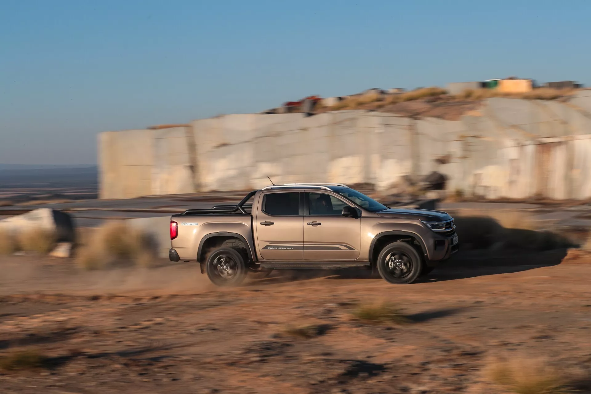 Dinamični bočni profilni pogled na pickup VW-Nutzfahrzeuge Amarok 2023 (2. generacija) v gibanju. Tovornjak je zajet s tehniko panoramskega fotografiranja, ostro izostren proti zamegljenemu ozadju, kar poudarja gibanje in hitrost. Ima kabino z dvema vrstama sedežev s štirimi vrati in polnimi zadnjimi sedeži, prikazuje agresivne linije karoserije, sodoben dizajn koles, stranska ogledala, ročaje vrat in tovornjakovo posteljo ob naravni nizki sončni svetlobi. To nakazuje na uporabo na prostem in v zahtevnem terenu ter potencialne sodobne avtomobilske tehnologije.